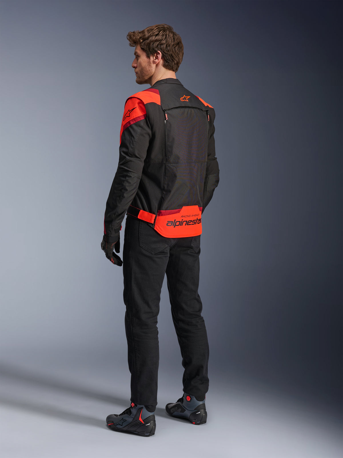 Mann in Alpinestars T-Stunt Air Jacke, Rückansicht, Schwarz, Fluo-Rot und Dunkelrot, Textil-Motorradjacke mit Mesh-Einsätzen und sportlichem Design, markantes Alpinestars-Branding auf dem unteren Rücken, kombiniert mit schwarzer Hose und Motorradstiefeln