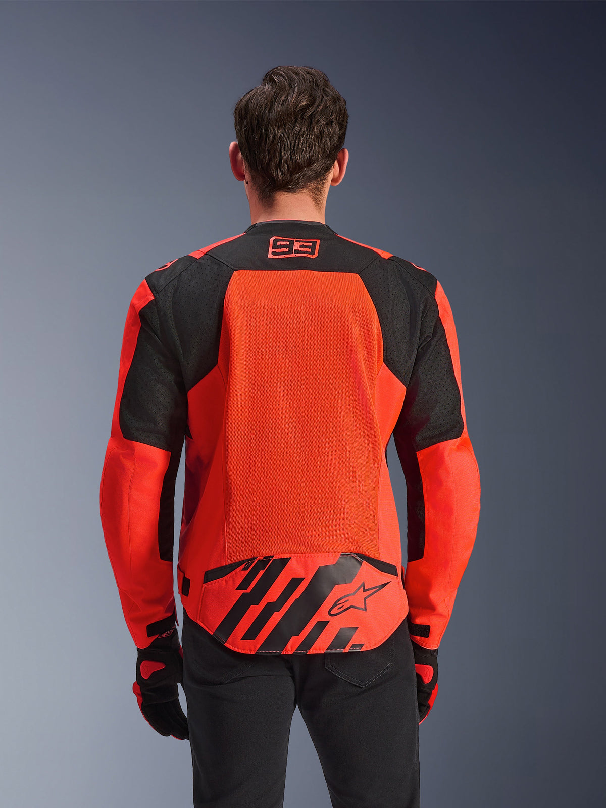 Alpinestars MM93 T-SPS Air V2 Jacke, Rückansicht, Rot Fluo und Schwarz, mit 93-Racing-Logo am Nacken, großem Mesh-Panel am Rücken für Luftzirkulation, unterer Rückenpartie mit Alpinestars Stern-Logo und geometrischen Streifen, getragen mit schwarzer Hose und Motorradhandschuhen