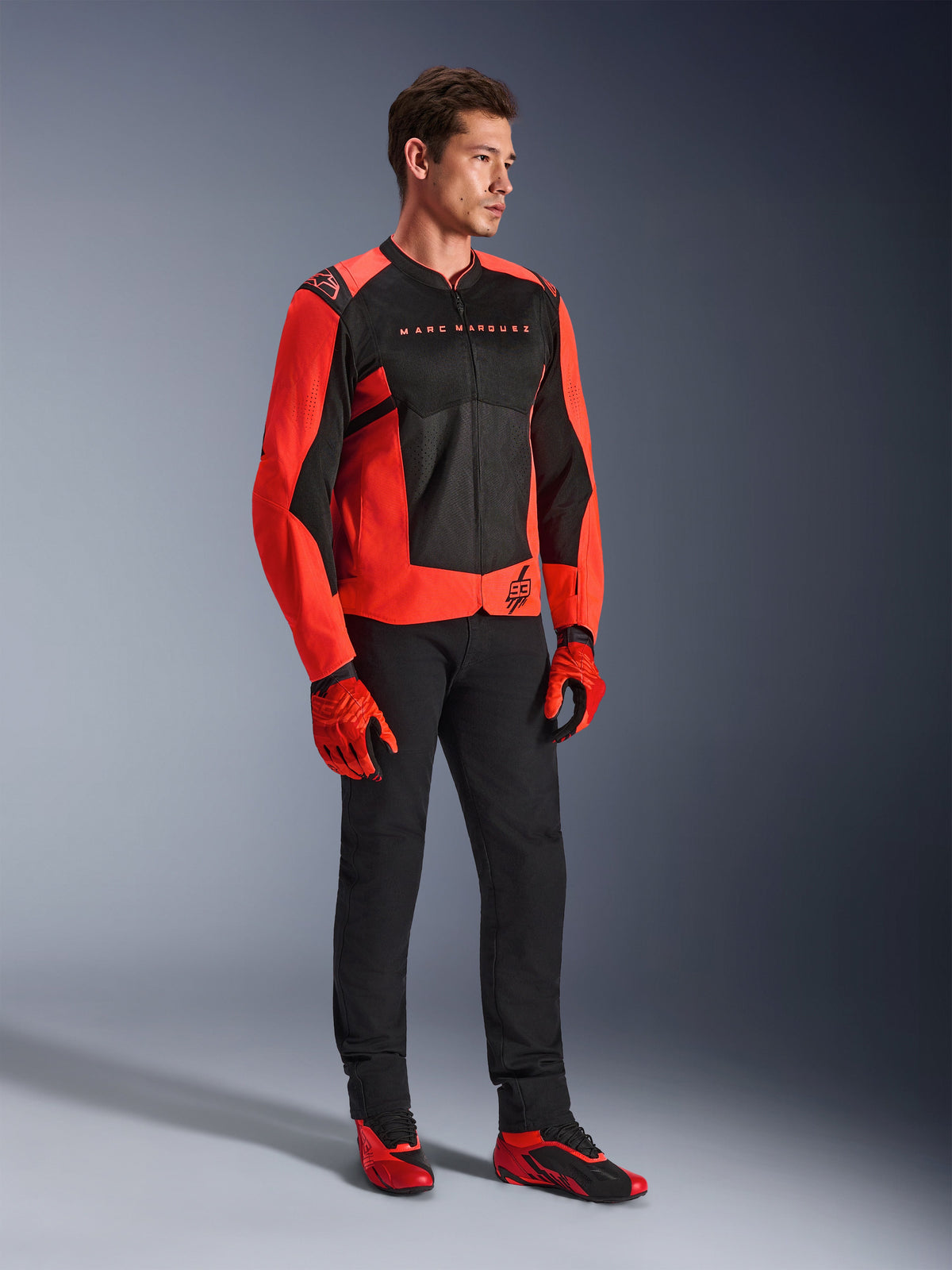 Alpinestars MM93 T-SPS Air V2 Jacke, Rot Fluo und Schwarz, getragen von einem Modell mit passender schwarzer Hose, rot-schwarzen Handschuhen und roten Motorradschuhen, mit Marc Marquez Branding und 93-Logo, sportorientierte urbane Fahrausrüstung