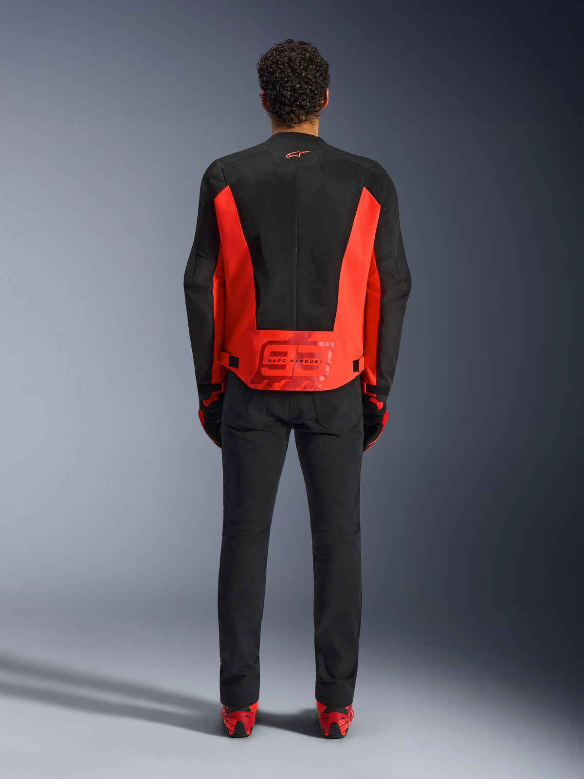 Alpinestars MM93 C-1 Air Jacke, Motorradjacke, Leuchtrot und Schwarz, Rückansicht mit Marc Marquez 93 Grafik am unteren Rücken, Tech-Mesh-Einsätze zur Belüftung, kombiniert mit schwarzer Hose und Ausrüstung mit roten Akzenten