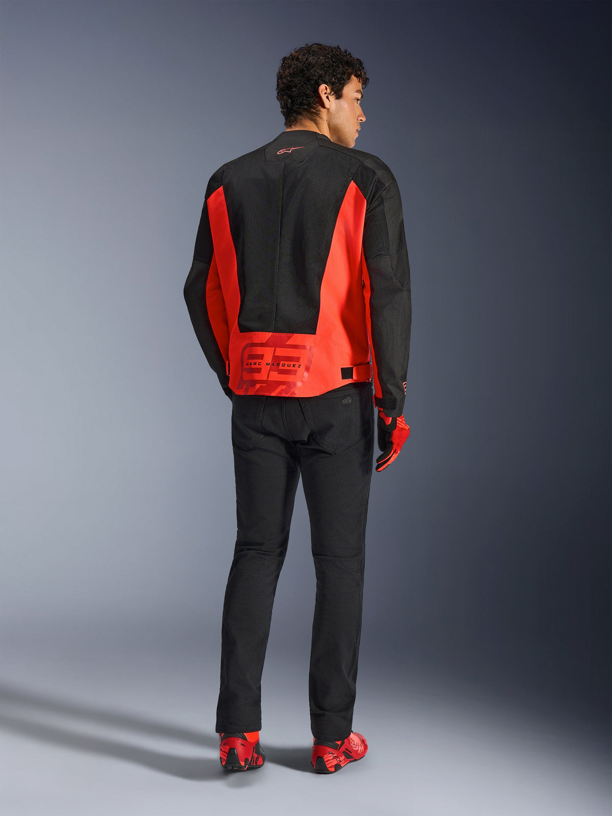 Rückansicht eines männlichen Models in der Alpinestars MM93 C-1 Air Jacke in Red Fluo und Schwarz, mit dem Marc Marquez 93 Logo auf dem unteren Rücken, kombiniert mit schwarzer Hose und roten Motorradschuhen, urbaner Motorrad-Pendler-Stil