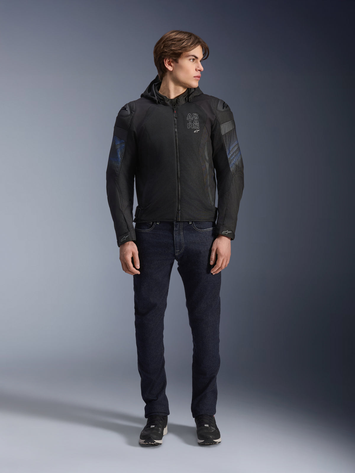 Männliches Model trägt die Alpinestars Zaca Air Venom WP Jacke in Schwarz und Electric Blue, Textil-Motorradjacke mit 3D-Mesh-Einsätzen und abnehmbarer Kapuze, kombiniert mit einer dunklen Denim-Jeans, moderne sportlich inspirierte Fahrausrüstung