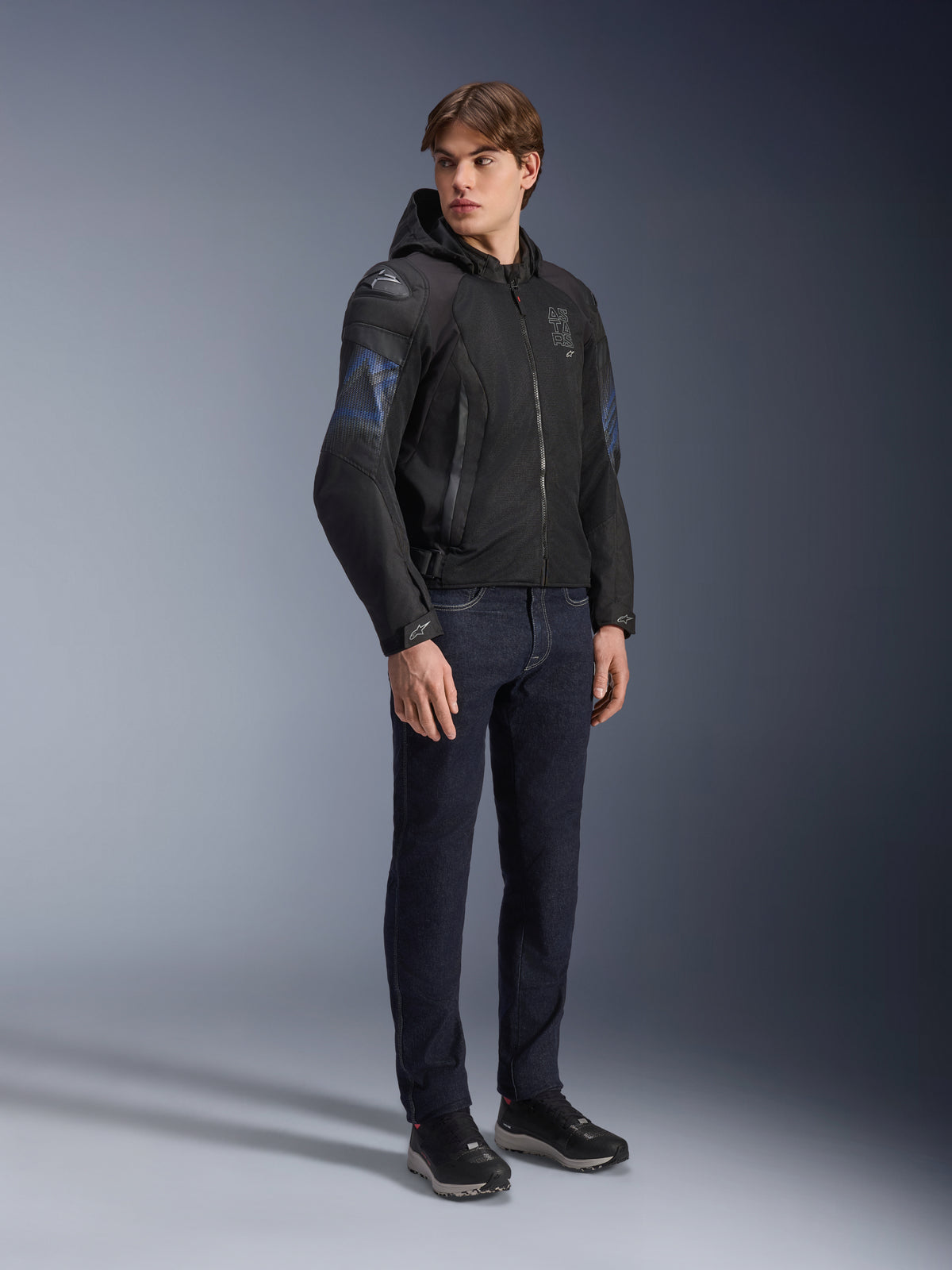 Alpinestars Zaca Air Venom WP Jacke, Textil-Motorradjacke in Schwarz und Electric Blue, getragen von einem Model mit dunkler Jeans und schwarzen Sneakern, mit abnehmbarer Kapuze, Mesh-Belüftungseinsätzen und DFS Lite Schulterschleifern, sportliche Allwetter-Fahrausrüstung.