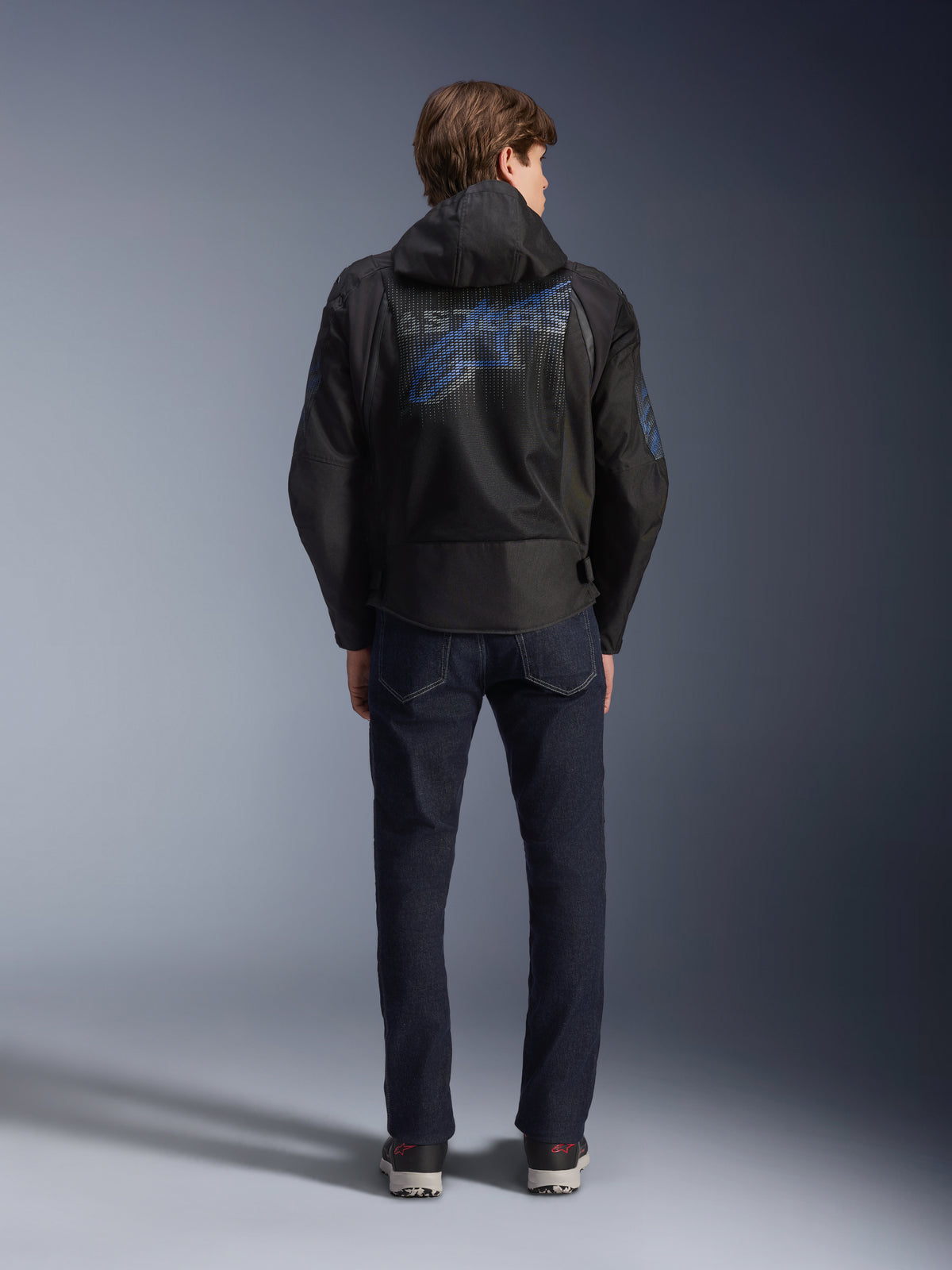 Alpinestars Zaca Air Venom WP Jacke, Schwarz und Electric Blue, Rückansicht mit einem großen blauen Alpinestars-Logo auf dem Mesh-Rückeneinsatz, abnehmbare Kapuze, getragen mit dunklen Denim-Jeans und Sneakern, sportliche Allwetter-Motorradausrüstung.