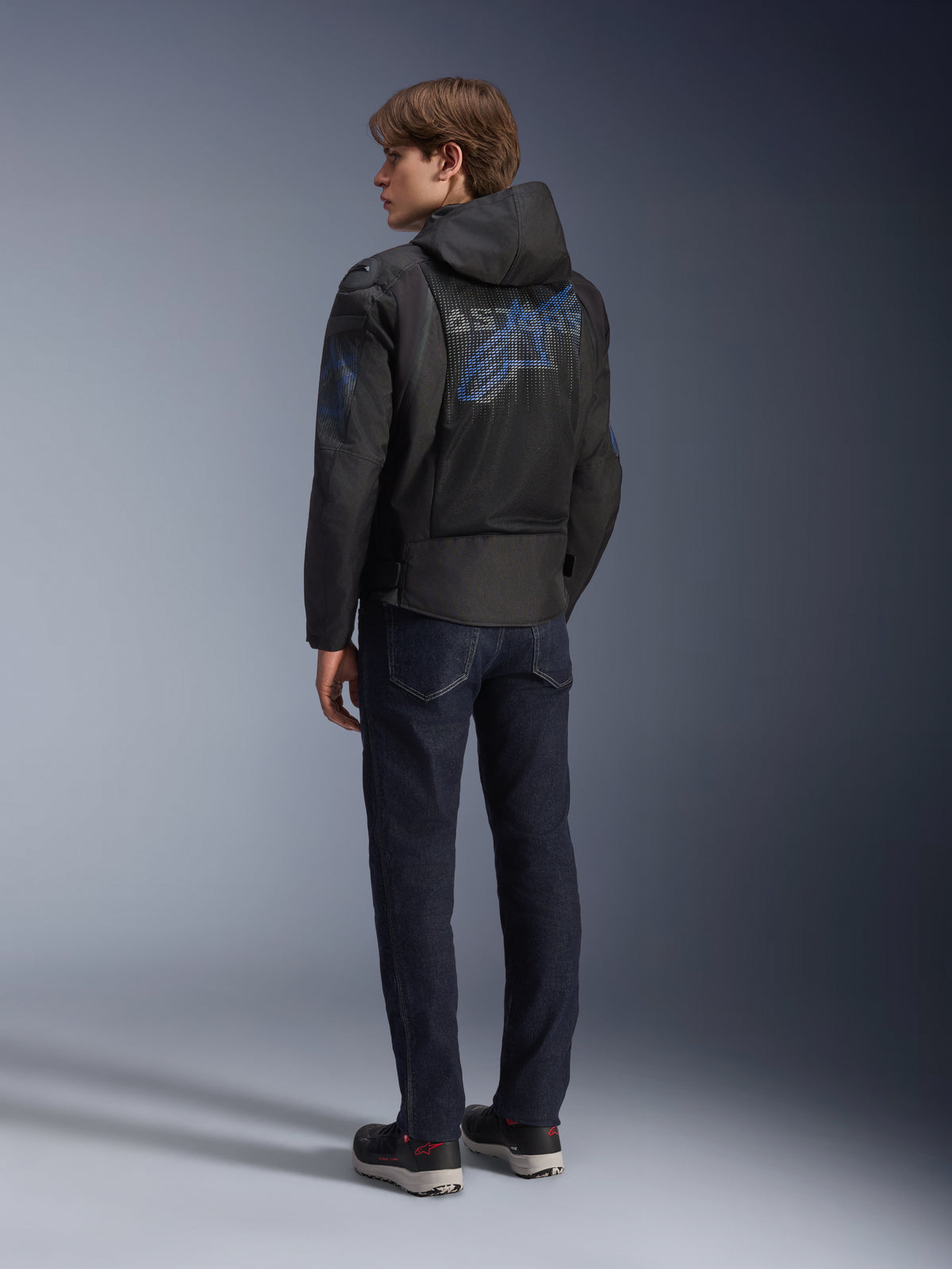 Alpinestars Zaca Air Venom WP Jacke, Schwarz und Electric Blue, Rückansicht an einem Modell, Mesh-Textil-Motorradjacke mit blauem Astar-Logo auf dem Rücken, mit abnehmbarer Kapuze, getragen mit dunkler Jeans und schwarzen Motorradschuhen.