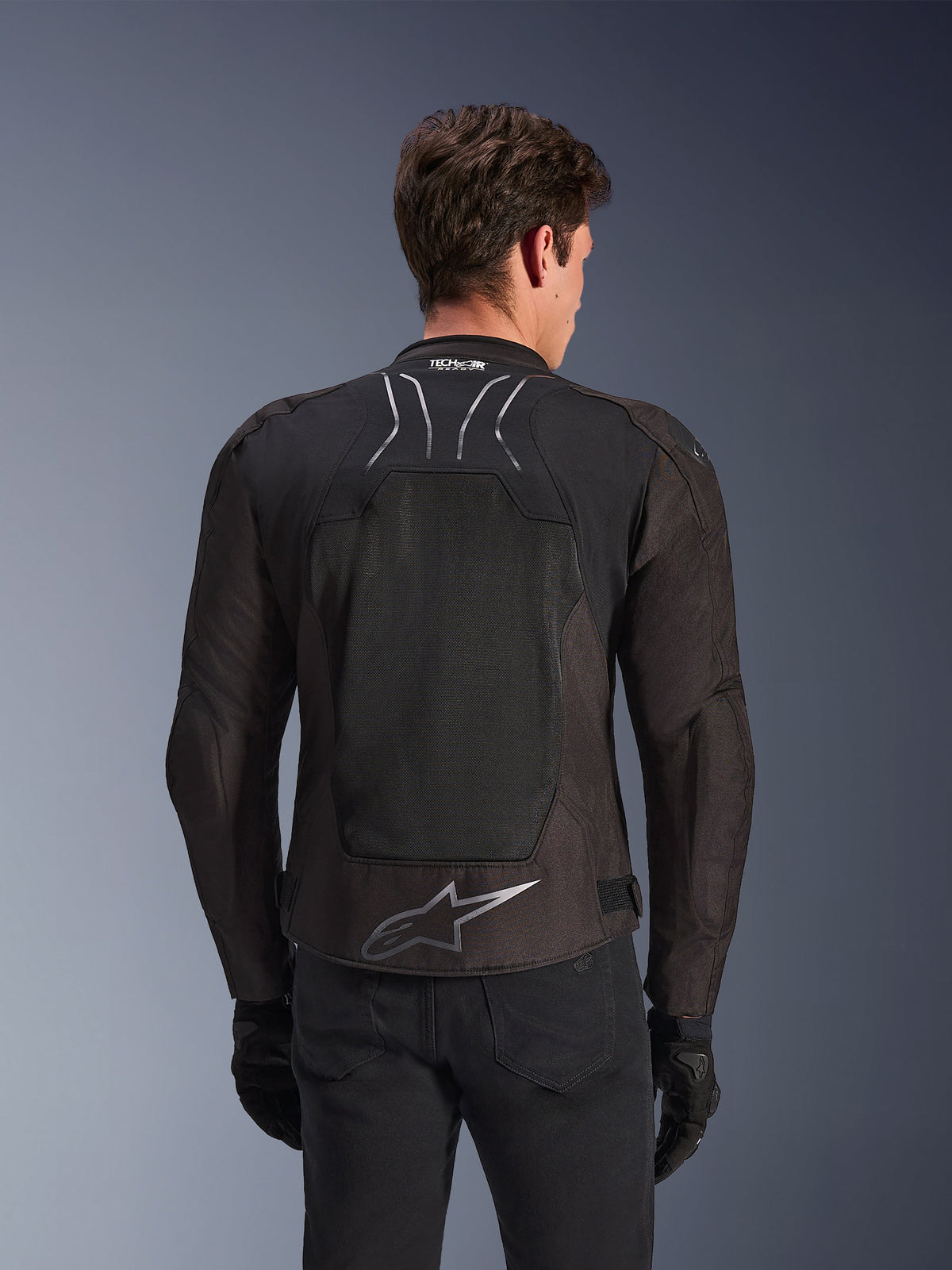 Alpinestars SMX Air Jacke in Black Anthracite, Rückansicht an einem Model, mit einem großen Tech-Mesh-Rückenpanel zur Belüftung, Tech-Air® Ready-Branding am Kragen und einem silbernen Astar-Logo am unteren Rücken, kombiniert mit schwarzen Handschuhen und einer Motorradhose.