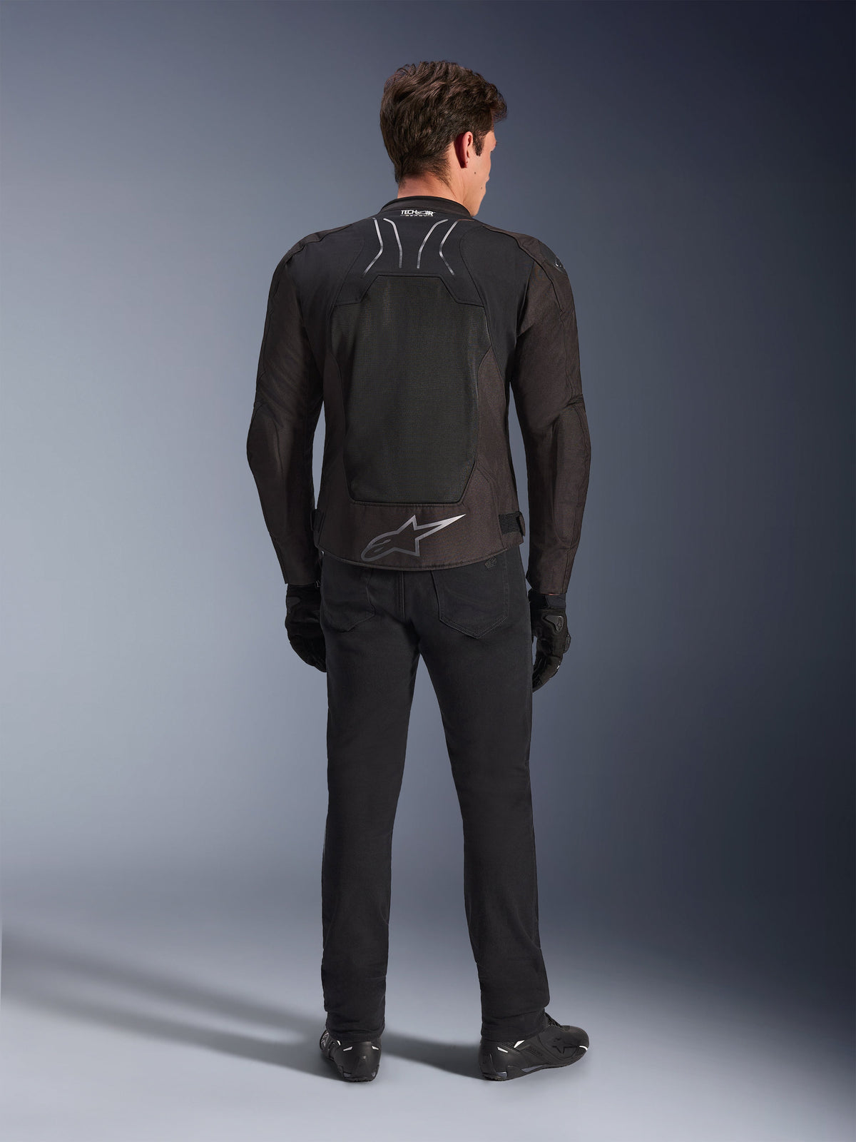 Alpinestars SMX Air Jacke, Schwarz Anthrazit, Rückansicht an männlichem Modell, mit großem silbernem Alpinestars-Logo am unteren Rücken und High-Flow-Mesh-Einsätzen, getragen mit schwarzer Motorradhose, Handschuhen und Stiefeln, sportliche Motorradausrüstung