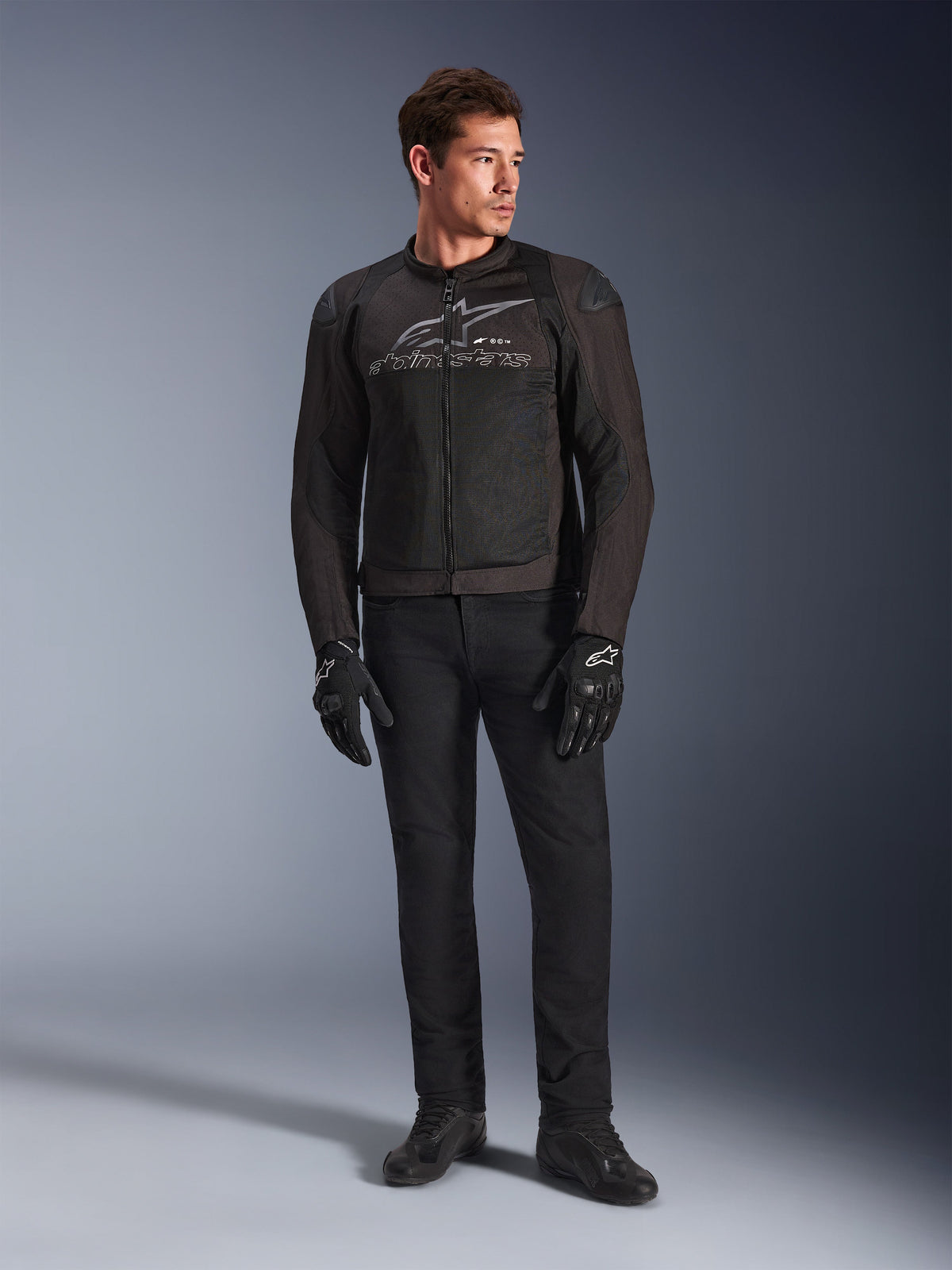 Männliches Model in der Alpinestars SMX Air Jacke, Motorradjacke in Black Anthracite mit belüfteten Mesh-Einsätzen und grauem Brust-Logo, kombiniert mit schwarzer Hose und Schutzhandschuhen, Studioaufnahme mit Farbverlauf im Hintergrund