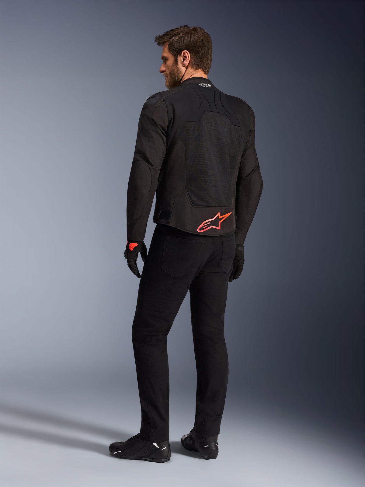 Rückansicht eines Modells in der Alpinestars SMX Air Jacke in Schwarz, mit einem leuchtend roten Alpinestars-Logo am unteren Rücken, „Tech-Air Ready“-Branding am Kragen und technischen Mesh-Einsätzen zur Belüftung, kombiniert mit schwarzer Motorradhose und Handschuhen.