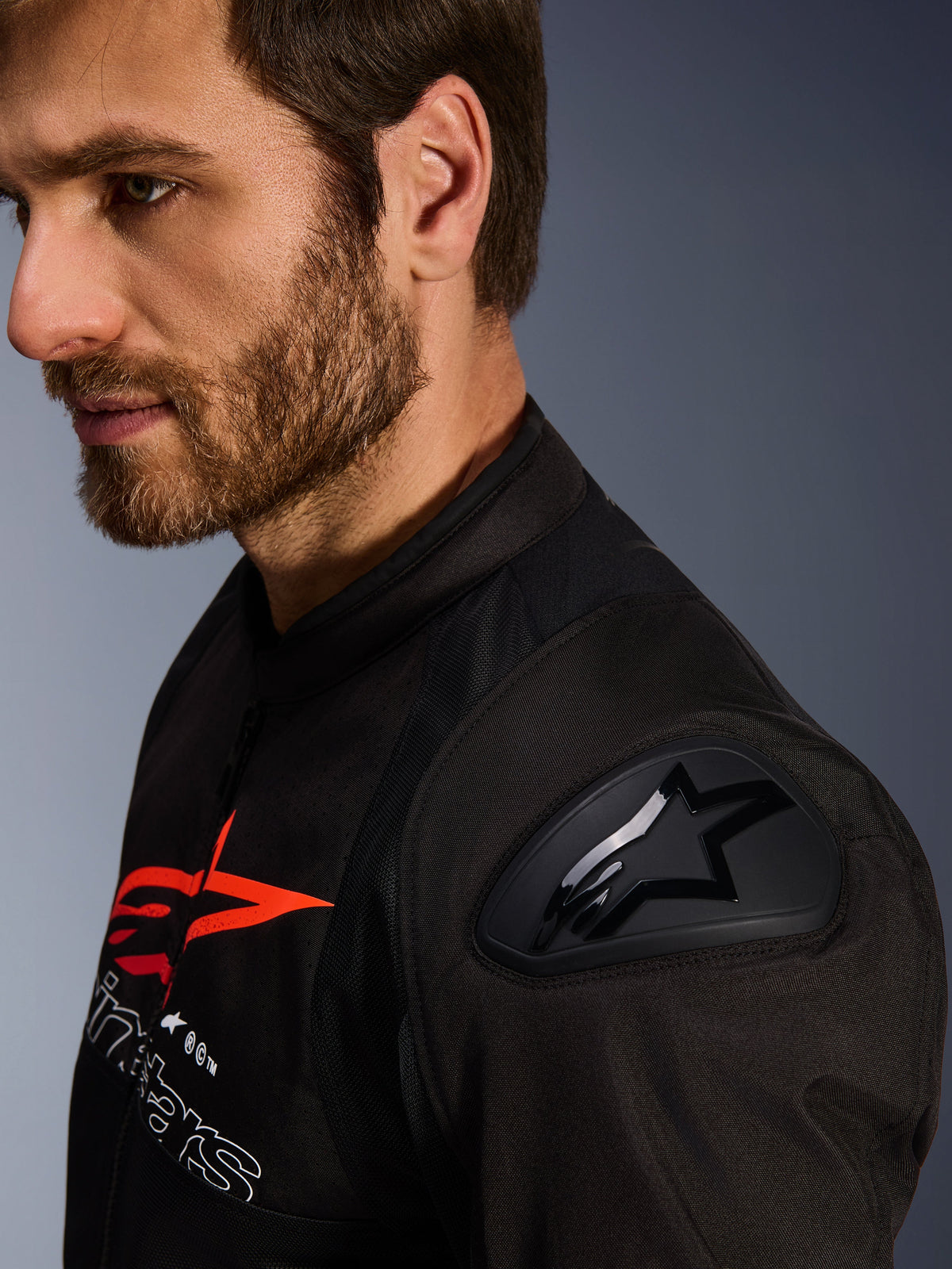 Alpinestars SMX Air Jacke, Schwarz und Rot, Nahaufnahme des DFS Lite Schulterprotektors mit Stern-Logo, getragen von einem bärtigen Mann, Textil-Renn- und Sport-Motorradjacke.