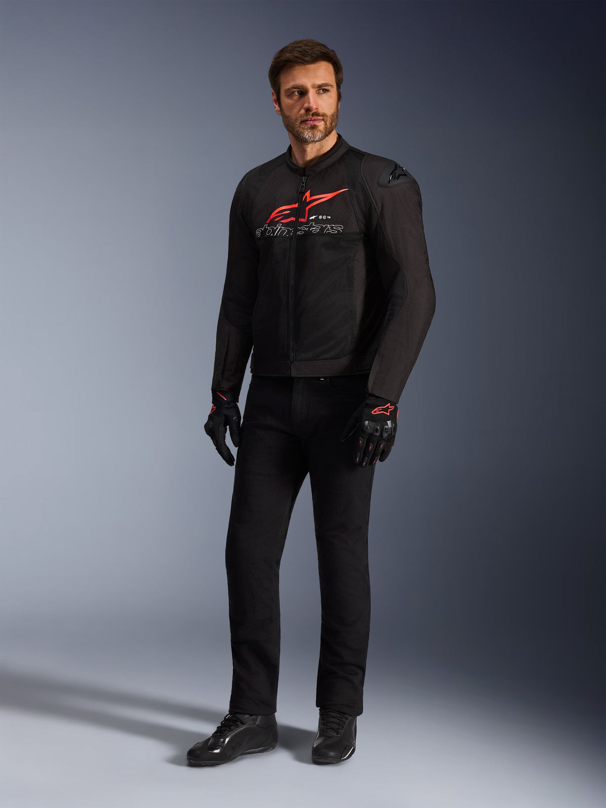 Männliches Model in der Alpinestars SMX Air Jacke in Schwarz Bright Red Weiß, kombiniert mit schwarzer technischer Hose, schwarz-roten Handschuhen und schwarzen Motorradschuhen, mit markantem rotem Brustlogo und weißem Branding, Ganzkörper-Studioaufnahme für Sport-Motorradausrüstung