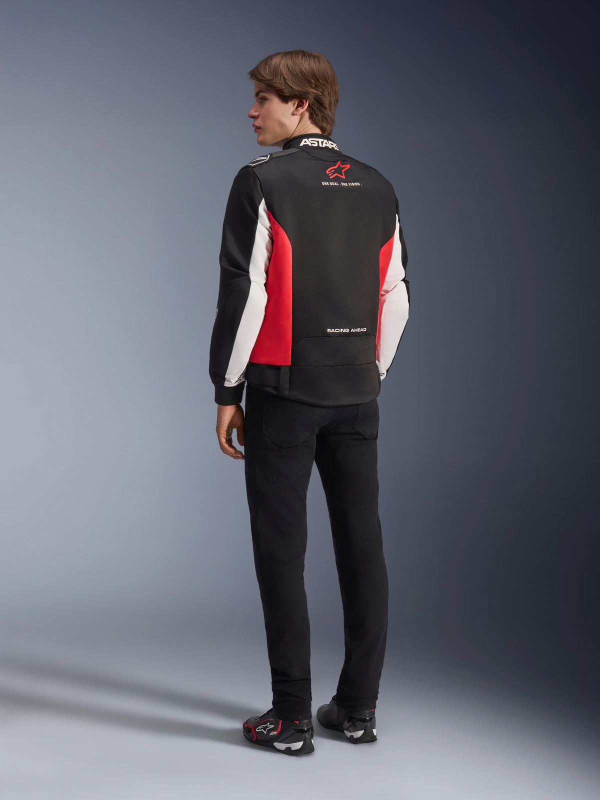 Alpinestars Monza Sport Jacke, Textil-Motorradjacke, Schwarz, Leuchtendes Rot und Weiß, Rückansicht des Modells mit roten Seiteneinsätzen und weißen Ärmelakzenten, Alpinestars Logo und 'ONE GOAL. ONE VISION.' Schriftzug, kombiniert mit schwarzer Hose und Motorradschuhen für einen sportlich inspirierten Look