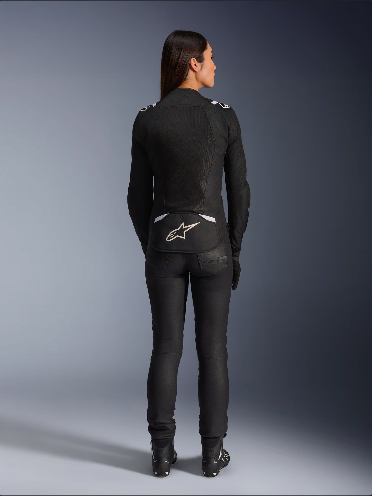 Alpinestars Stella T-SPS Air v2 Jacke, Damen-Motorradjacke, Schwarz und Weiß, Rückansicht mit Mesh-Belüftungseinsätzen und weißem Logo am unteren Rücken, getragen mit schwarzer Fahrhose und Stiefeln, urbane Sportausrüstung.