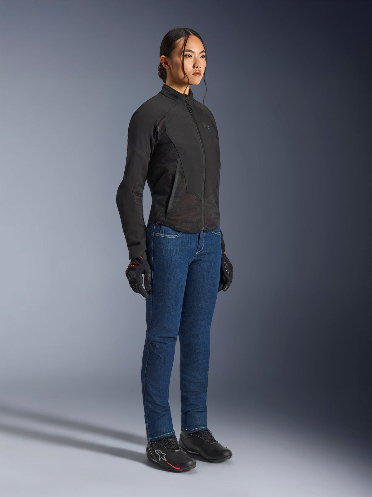 Female rider wearing the Alpinestars Stella Aeroshell Airflow Jacket in Black Anthracite, slim-fit softshell motorcycle jacket, paired with blue riding jeans and black gloves, full-body studio view