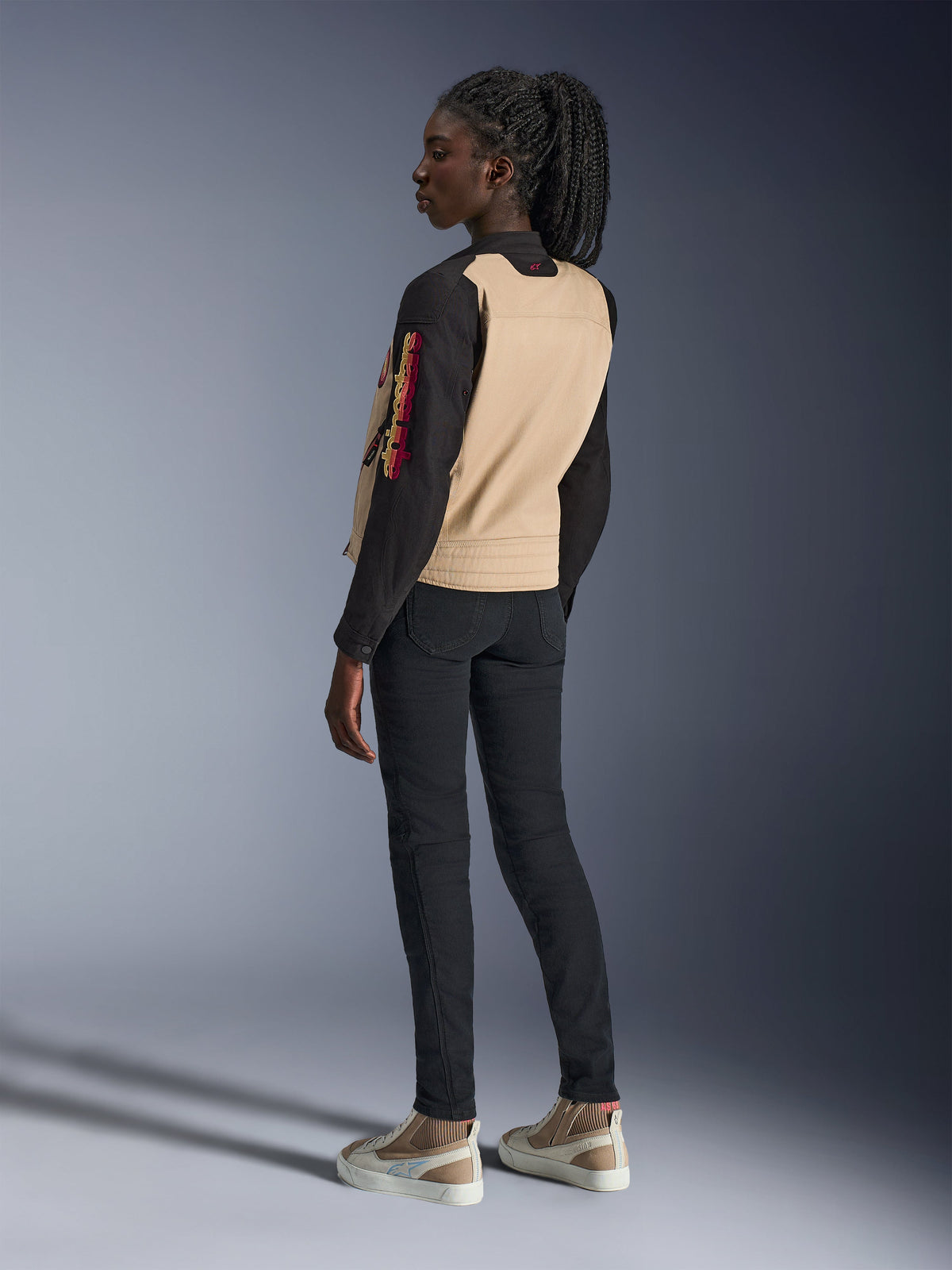 Alpinestars Stella Grace Damenjacke, urbane Motorradjacke, Aluminum Black mit hellbraunem Rückenteil und schwarzen Ärmeln, Rückseitenansicht an einem weiblichen Model, mit besticktem farbenfrohem Branding auf dem Ärmel und plissierter Schulterkonstruktion