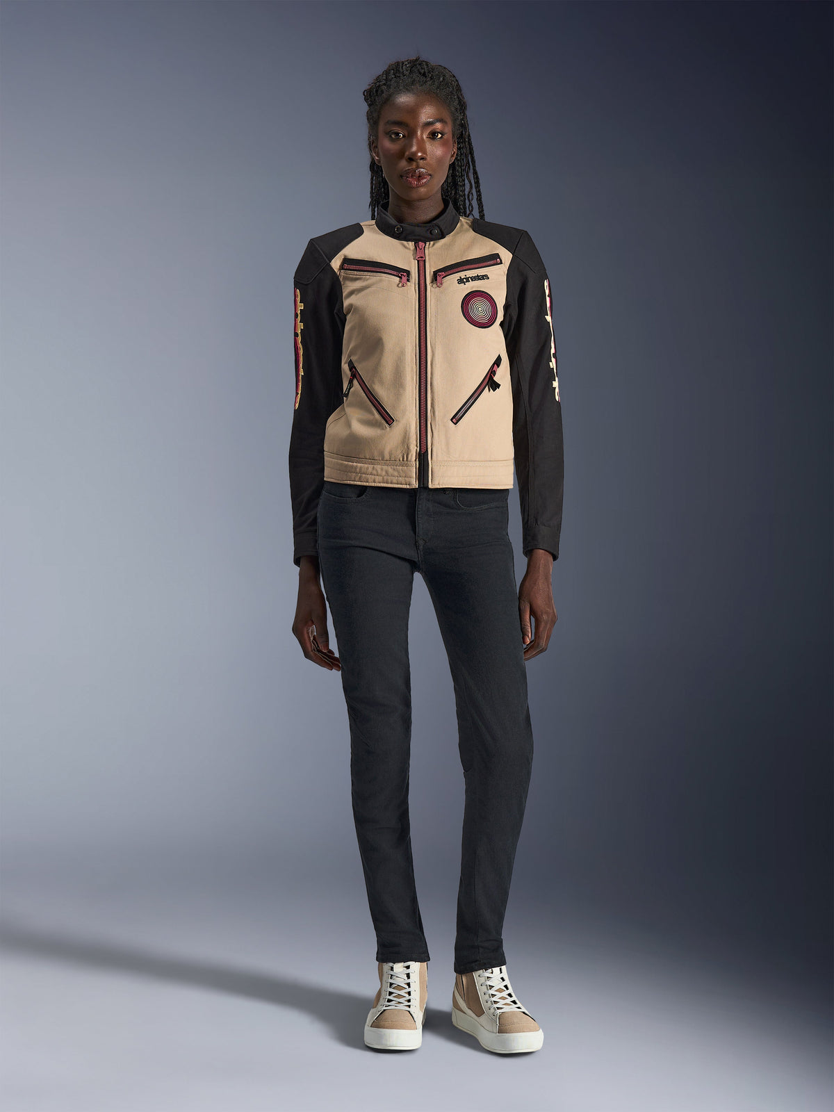 Alpinestars Stella Grace Damenjacke, urbane Motorradjacke, hellbraune und schwarze Textilkonstruktion, mit Reißverschlüssen vorne und besticktem Oscar-Spiral-Aufnäher, getragen mit schwarzer Hose und High-Top-Sneakern, moderne, von Workwear inspirierte Fahrausrüstung