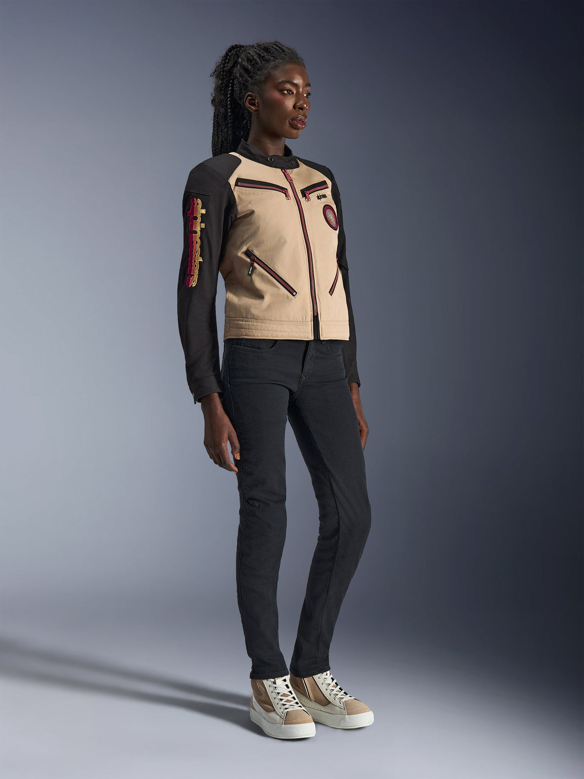 Model trägt die Alpinestars Stella Grace Jacke in Aluminum Black, mit beigefarbenem Rumpf und schwarzen Ärmeln mit roten Reißverschluss-Akzenten, kombiniert mit einer schmal geschnittenen schwarzen Hose und cremefarbenen Sneakern, moderne urbane Motorradbekleidung für Damen