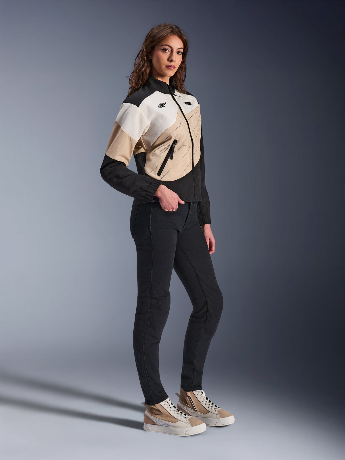 Alpinestars Stella Moshell Jacke, urbane Motorradjacke für Damen, Black Sand und Off White, mit asymmetrischem geometrischem Design, getragen mit schmaler schwarzer Hose und hellbraunen Sneakern, Ganzkörper-Studioaufnahme für moderne urbane Fahrausrüstung.