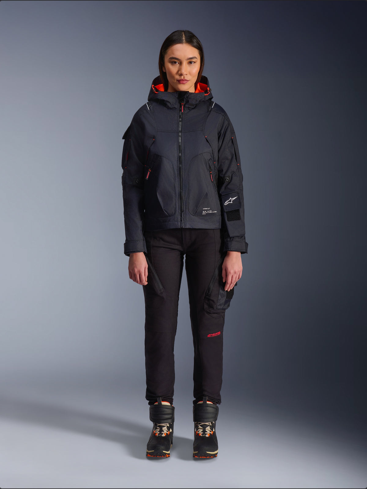 Alpinestars Actea Damenhose, schwarze Motorradhose für Damen mit roten Schriftelementen und Cargotaschen, getragen von einer Fahrerin mit schwarzer Kapuzenjacke, moderne urbane Motorradbekleidung