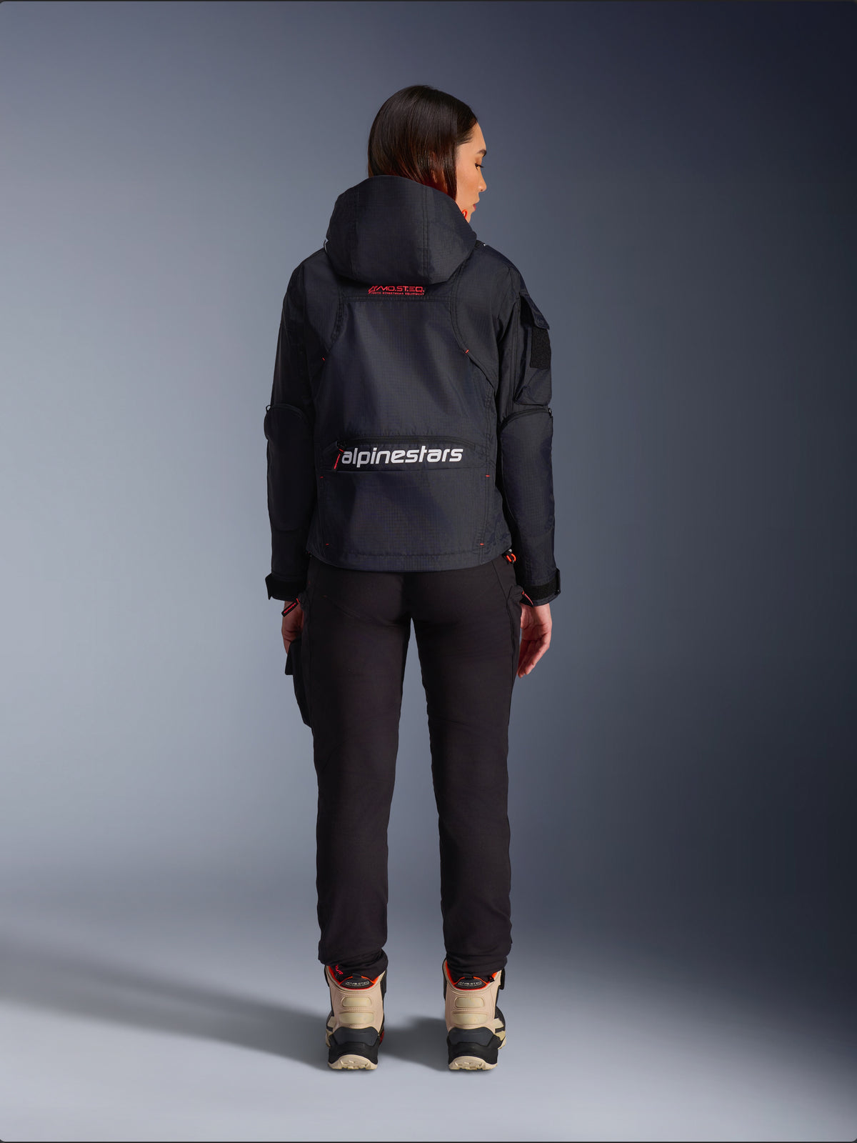 Alpinestars Actea Damen-Hose, schwarze Motorradhose mit verjüngter Passform, kombiniert mit einer dunklen Molly Air Jacke mit Kapuze und beigefarbenen Stiefeln, Rückansicht einer Fahrerin in urbaner Motorradausrüstung