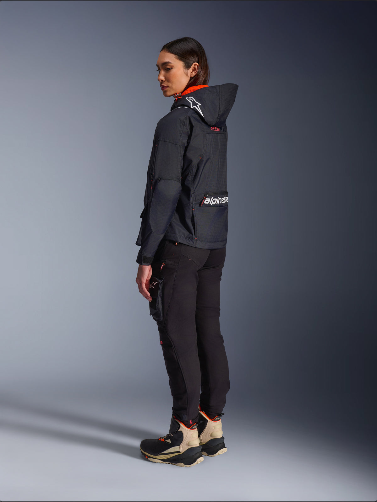 Alpinestars Actea Damen-Hose, Schwarz, moderne Motorradhose mit schmal zulaufender Passform und Cargo-Tasche am Oberschenkel, Seitenansicht von hinten an einem Modell mit dunkler Kapuzenjacke und Motorradstiefeln, urbaner Fahrstil.