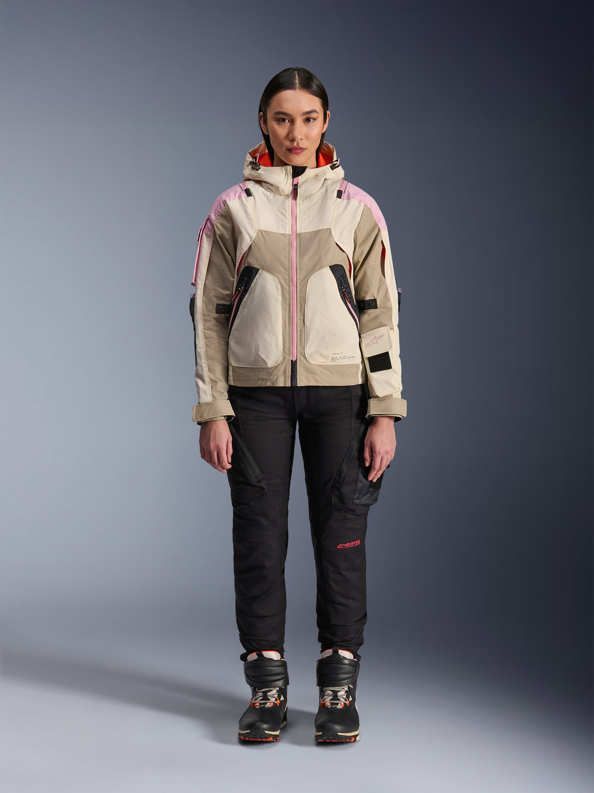Weibliches Model in der Alpinestars Molly Air Jacke in Sand Pink Almond, kombiniert mit einer schwarzen technischen Motorradhose und Stiefeln, Ganzkörperansicht von vorne, urbane Motorradjacke mit Utility-inspiriertem Design und rosa Akzenten