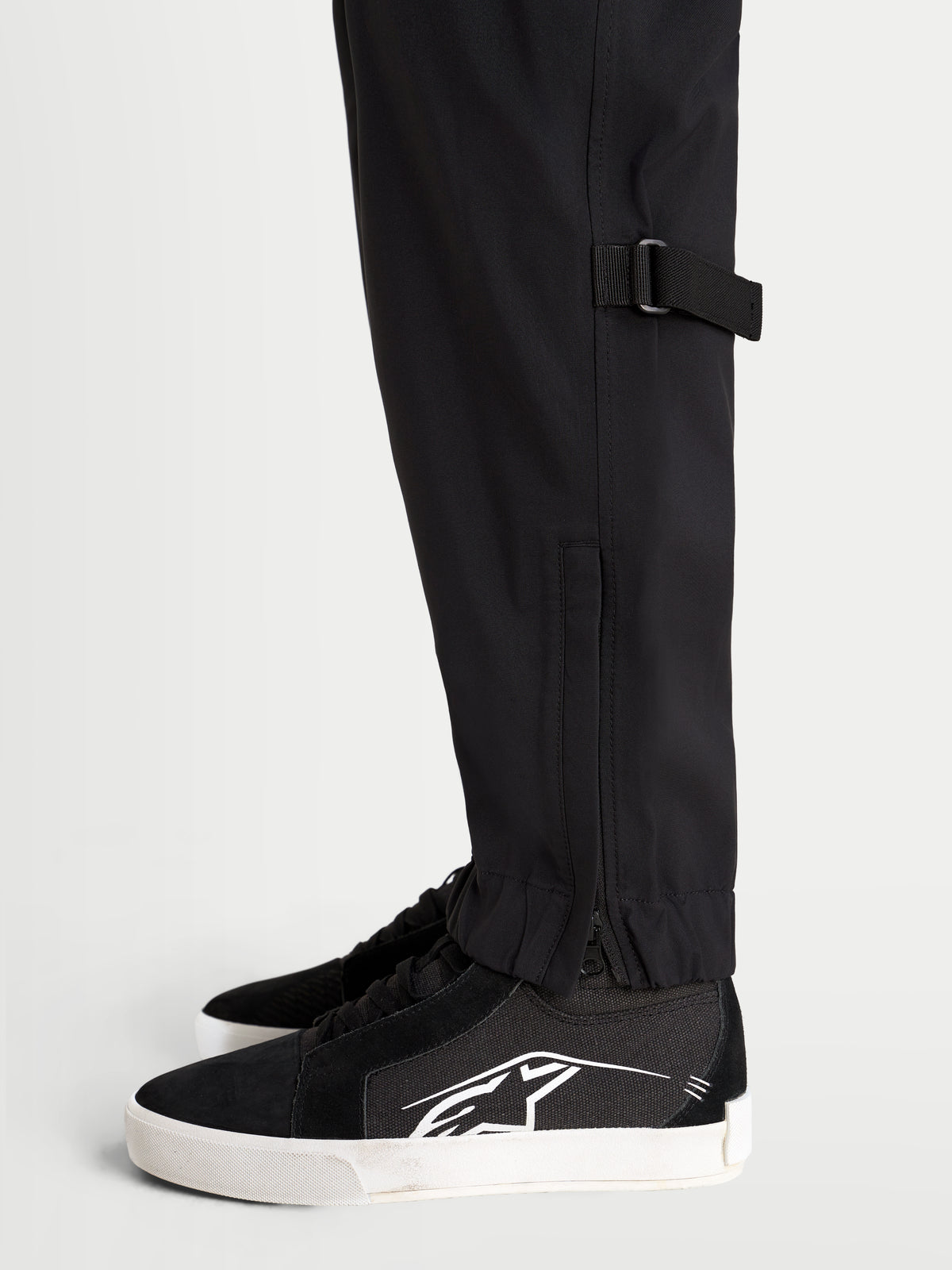 Alpinestars Juggernaut V 2 Drystar Hose, urbane wasserdichte Herren-Motorradhose in Schwarz, Nahaufnahme der Seitenansicht mit Unterschenkelverstellung und elastischem Saum mit Reißverschlussöffnung an Model