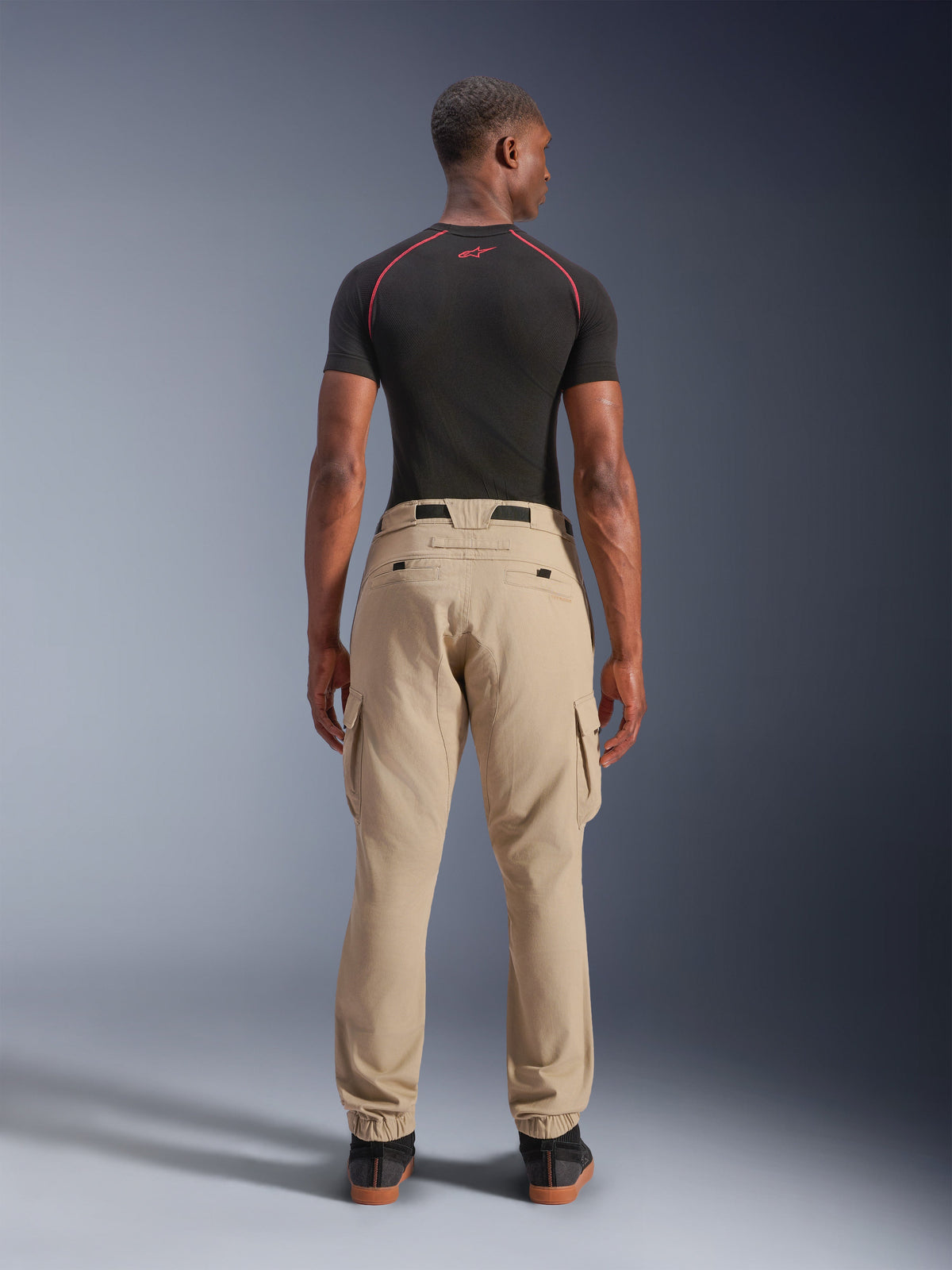 Alpinestars Flex-AST Cargo-Hose – Kurz, Hellbeige, Rückansicht an einem Model, reguläre Tapered-Passform mit elastischen Säumen und Cargotaschen, getragen mit einem schwarzen technischen Base Layer mit roten Akzenten, moderne urbane Motorradbekleidung
