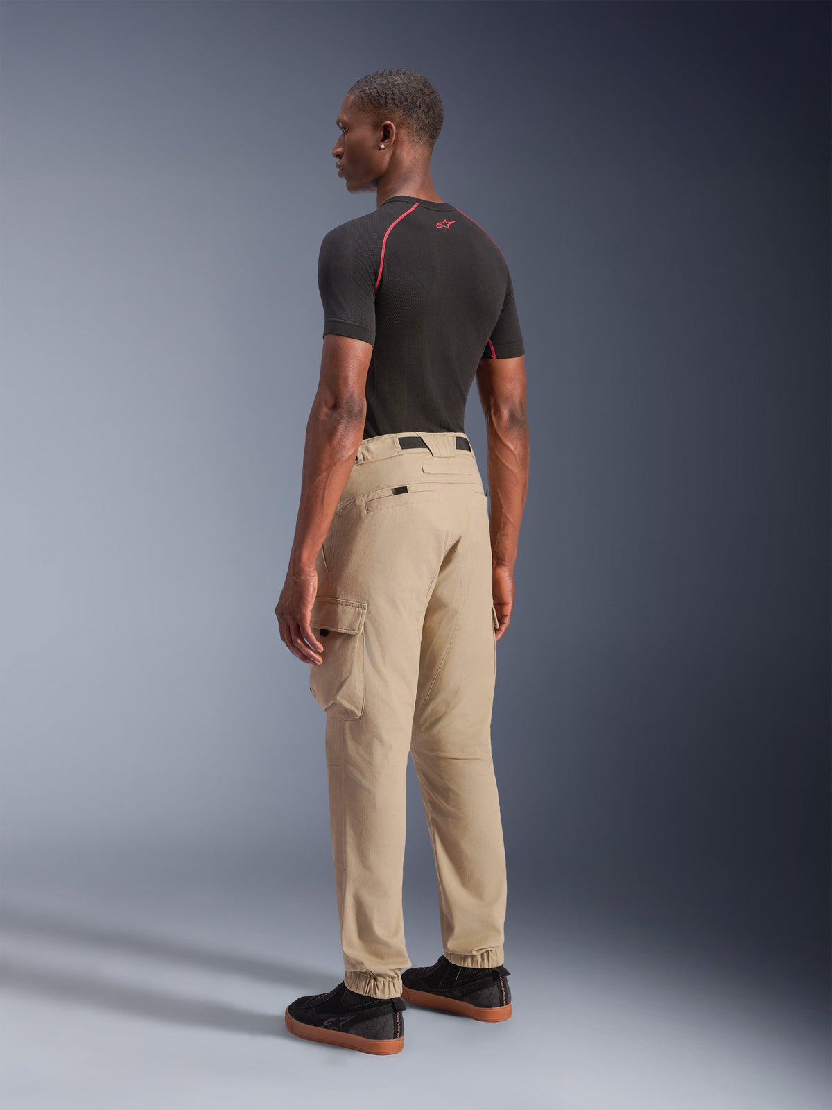 Alpinestars Flex-AST Cargo-Hose, urbane Motorradhose, Hellbeige, von einem Model in einer rückseitigen Seitenansicht getragen, mit funktionalen Cargotaschen und elastischen Bündchen, kombiniert mit einem schwarzen Kurzarm-Shirt und schwarzen Sneakern.