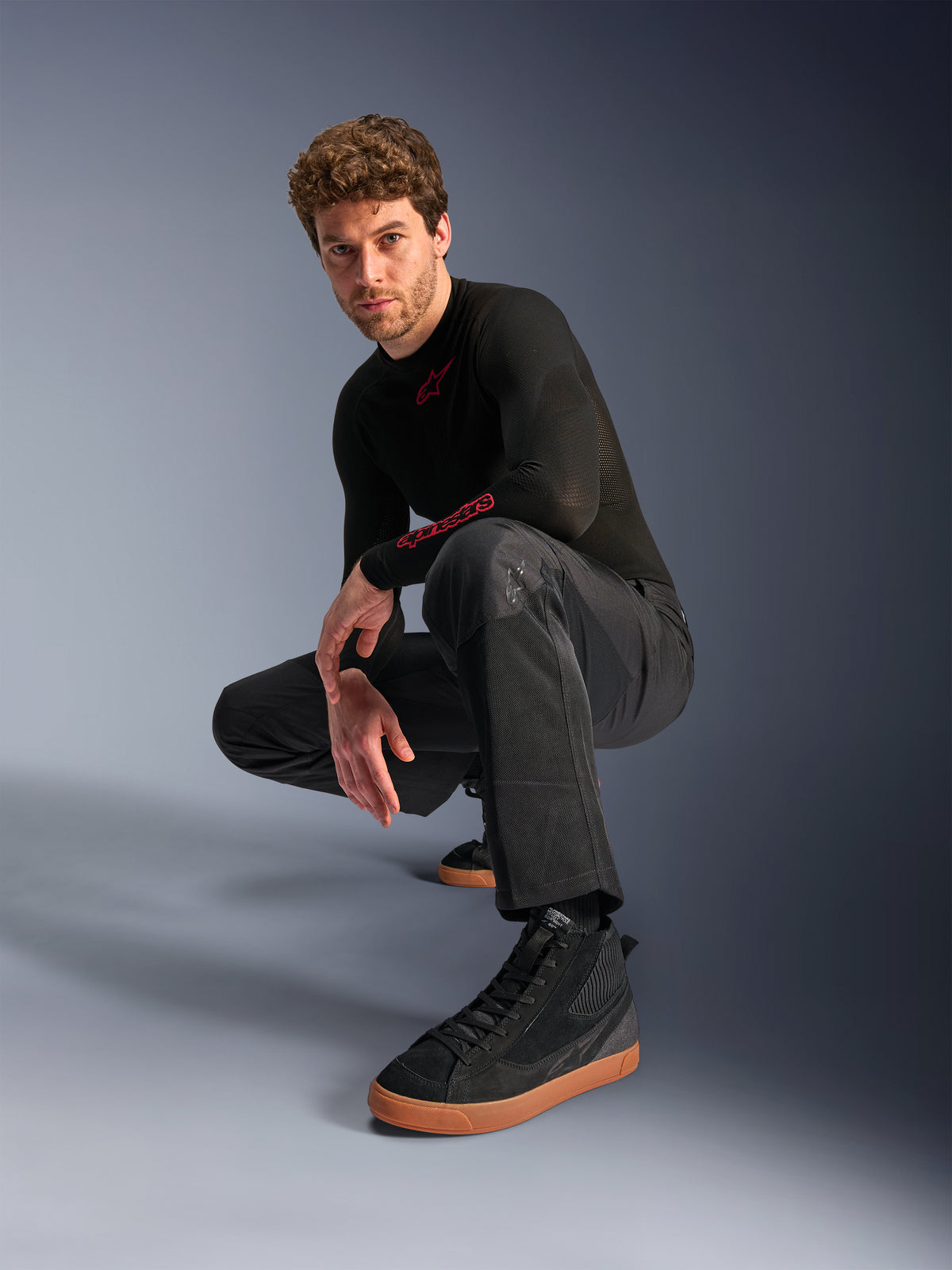 Alpinestars C-1 Air Hose, Urbane Motorradhose, Schwarz, Modell hockend in einer Studio-Umgebung, kombiniert mit schwarzem Langarm-Oberteil und schwarzen Sneakern mit hellbraunen Sohlen, mit atmungsaktiven Mesh-Einsätzen und modernem urbanen Stil