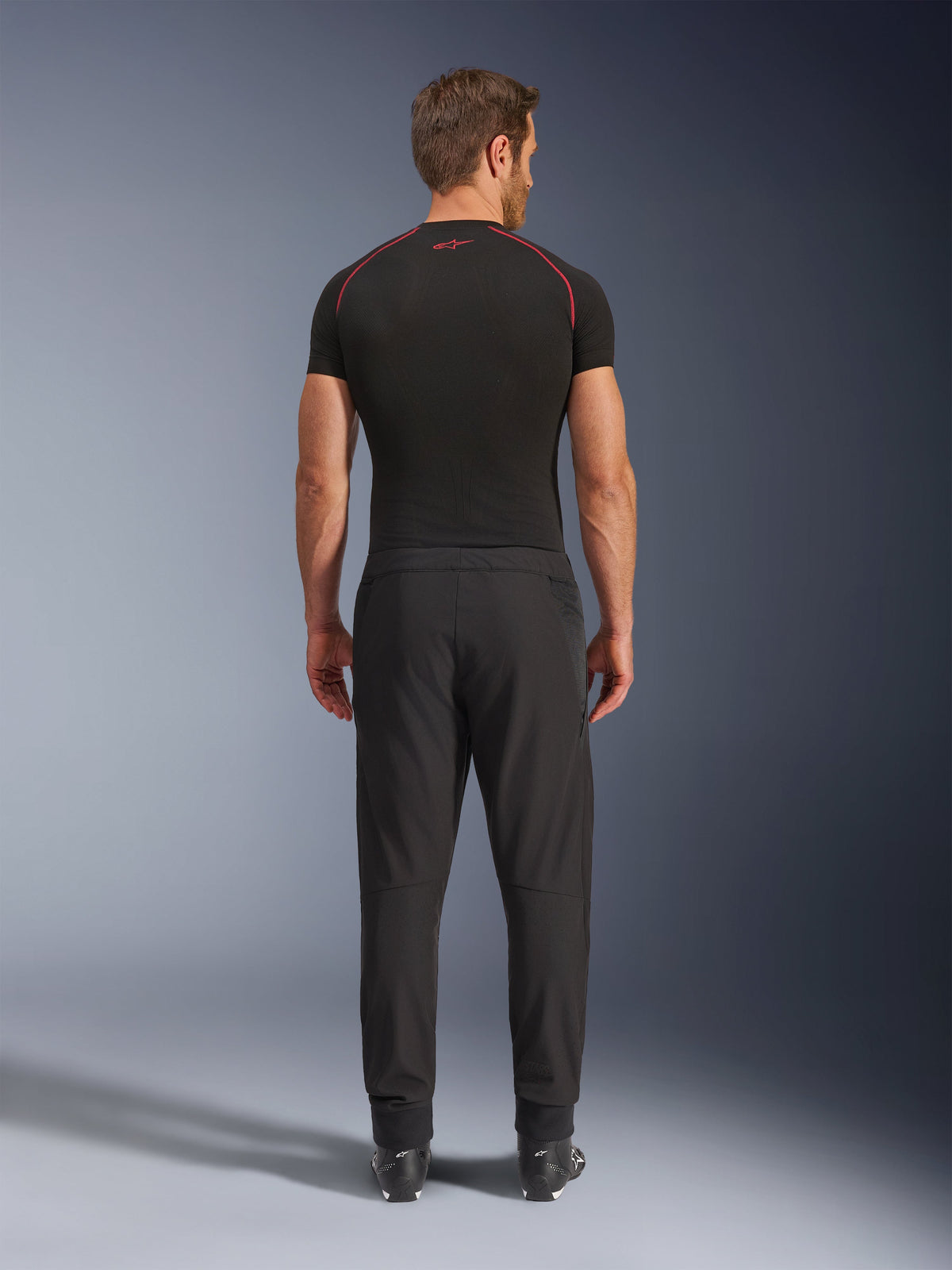Alpinestars Aeroshell Airflow Hose, schwarze Motorradhose, Rückansicht an einem Model mit schwarzem Baselayer und Stiefeln, mit einem leichten Softshell-Chassis und integrierten Mesh-Einsätzen zur Belüftung.