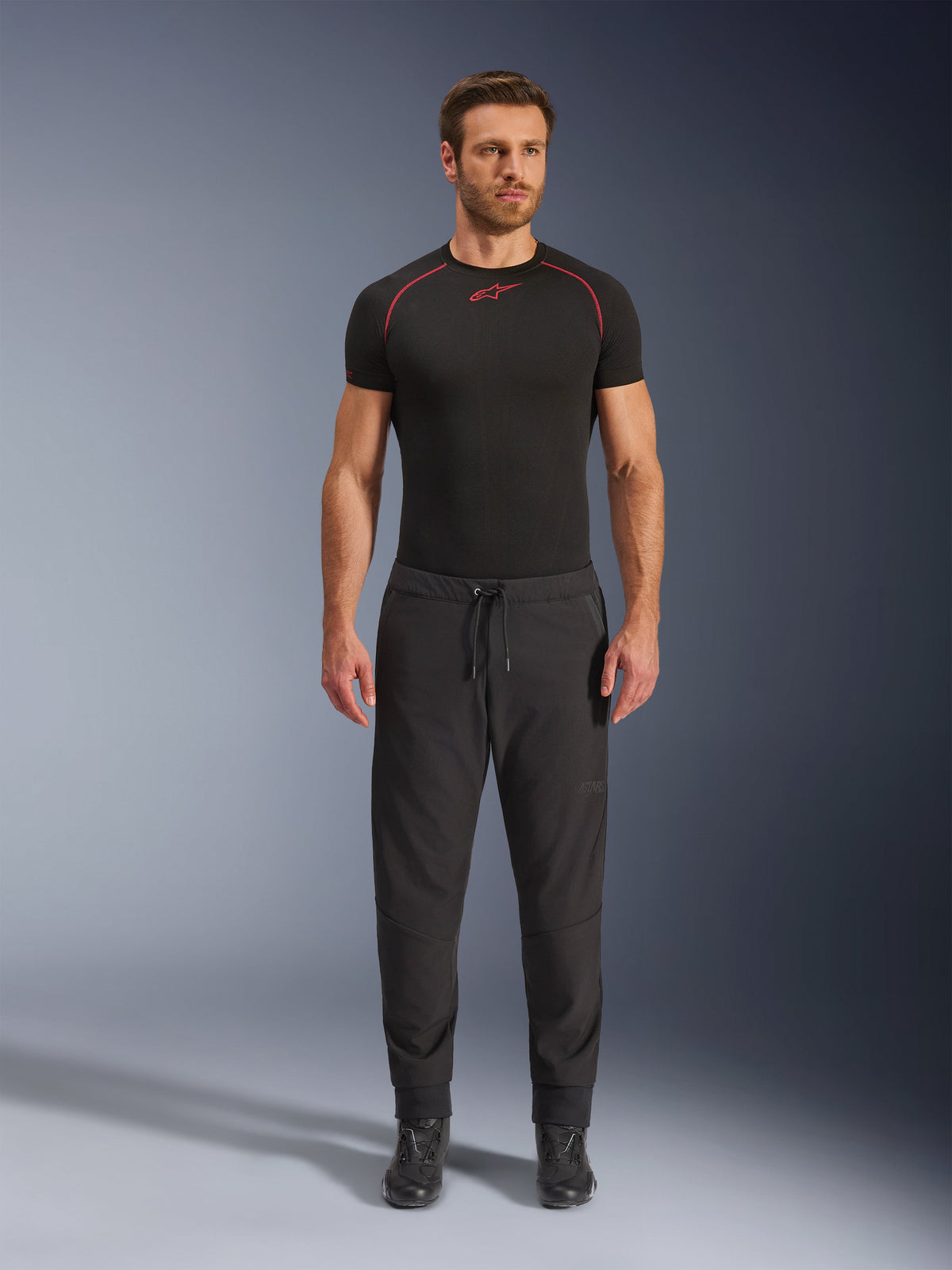 Model trägt die Alpinestars Aeroshell Airflow Hose in Schwarz, kombiniert mit einem schwarzen kurzärmeligen Baselayer und schwarzen Motorradstiefeln. Diese leichte Softshell-Motorradhose verfügt über einen Kordelzug am Bund sowie elastische Bündchen und ist für Sport- und Tourenfahrer bei warmen Wetterbedingungen konzipiert.