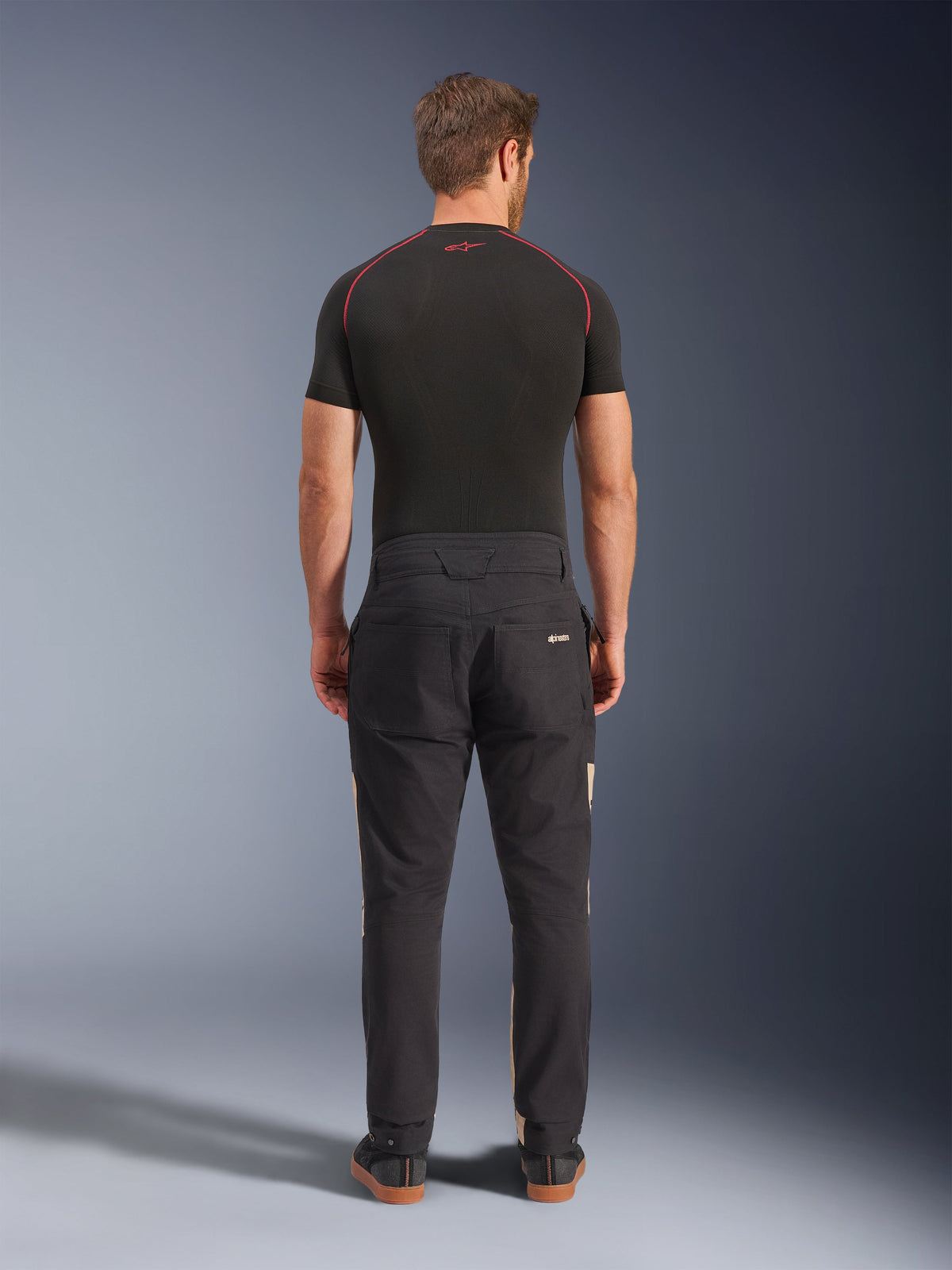 Alpinestars Flex-AST Canvas Pants in Black Aluminum, rear view on a model, featuring workwear-inspired styling with back pockets and subtle tan side panels, worn with a black performance base layer and casual riding sneakers.