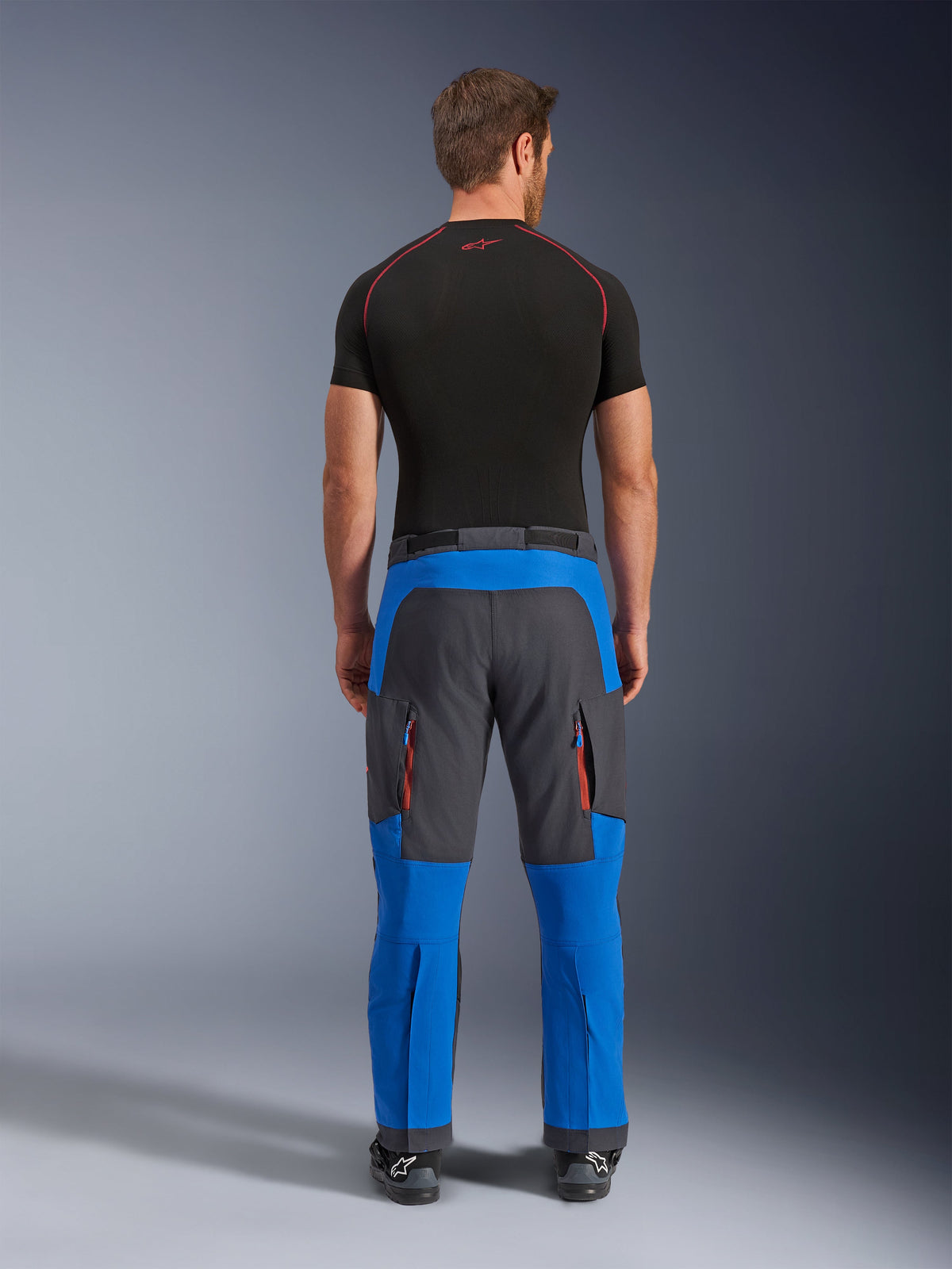 Alpinestars Maxdura Dual Hose in Royal Blue und Deep Gray, Adventure-Motorradhose in der Rückansicht, mit Cargo-Reißverschlusstaschen mit orangefarbenen Akzenten und Über-Stiefel-Design, getragen mit einem schwarzen kurzärmeligen Baselayer