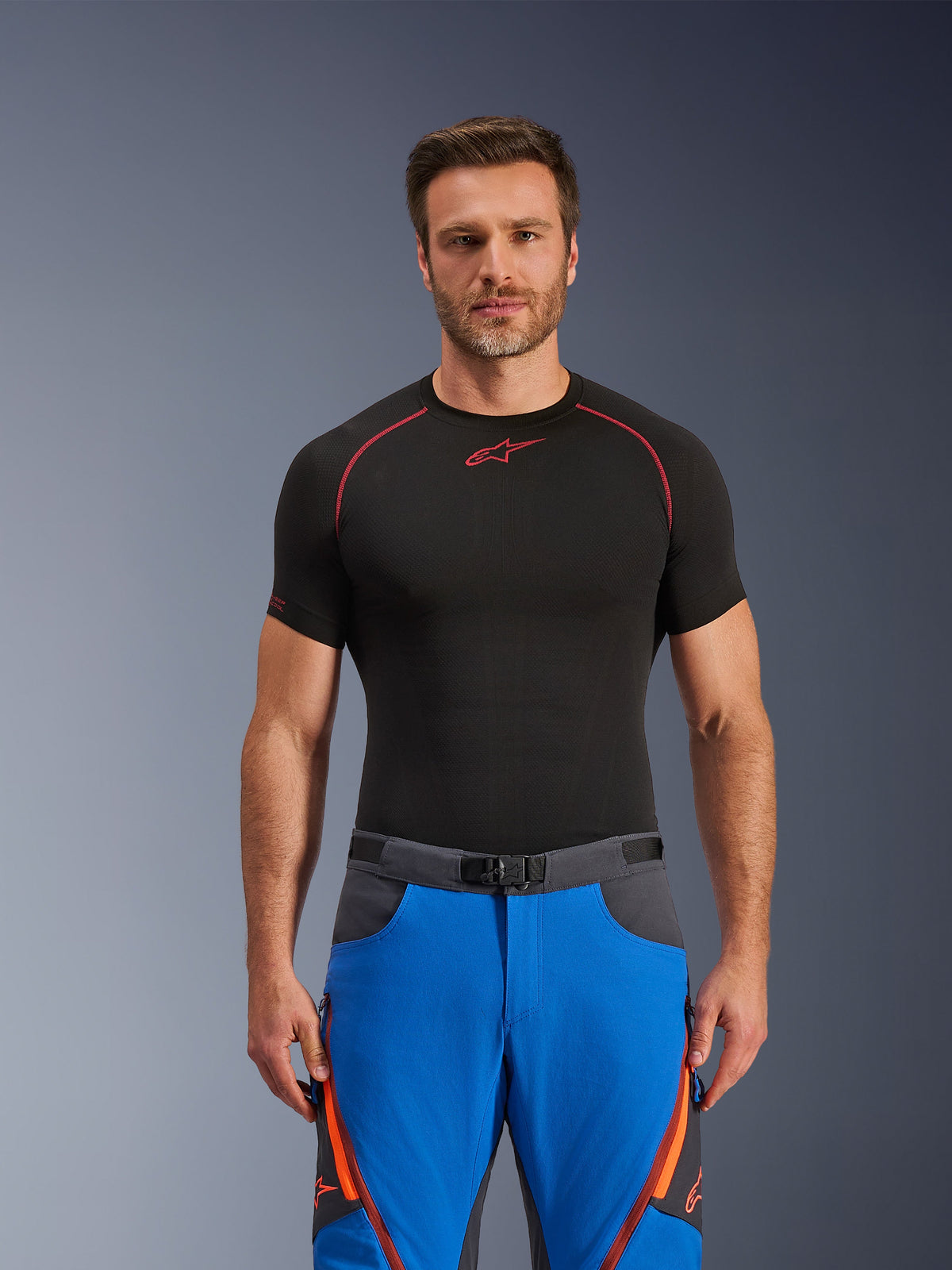 Alpinestars Maxdura Dual Hose, Königsblau und Dunkelgrau, Adventure-Motorradhose mit orangefarbenen Akzenten, getragen von einem Model mit schwarzem Kompressionsshirt, mit Magnetschnallen-Gürtel und Belüftungsreißverschlüssen am Oberschenkel, professionelle Motorsportbekleidung