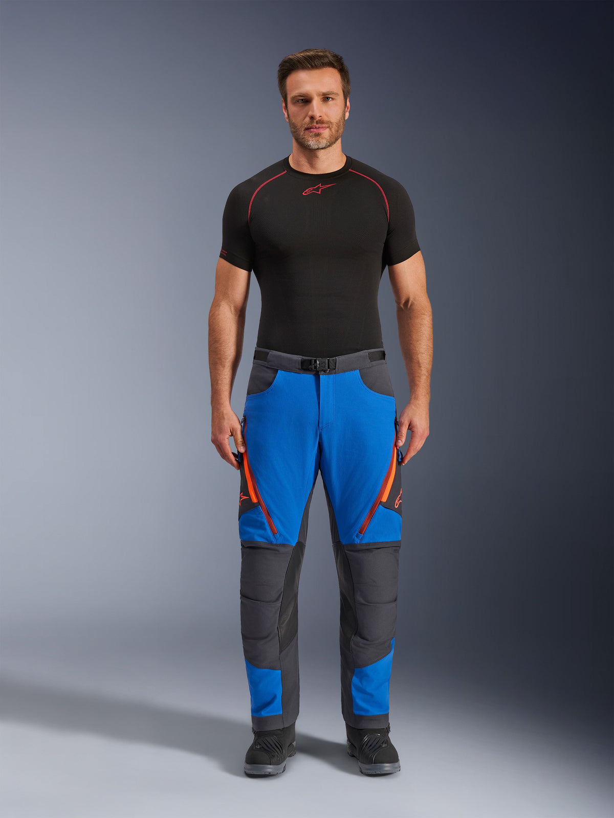 Alpinestars Maxdura Dual Hose, Adventure-Motorradhose, Royalblau und Dunkelgrau, orangefarbene Reißverschluss-Akzente, getragen mit einem schwarzen Alpinestars Base Layer und schwarzen Stiefeln, mit verstärkten Knieeinsätzen und technischem Offroad-Design