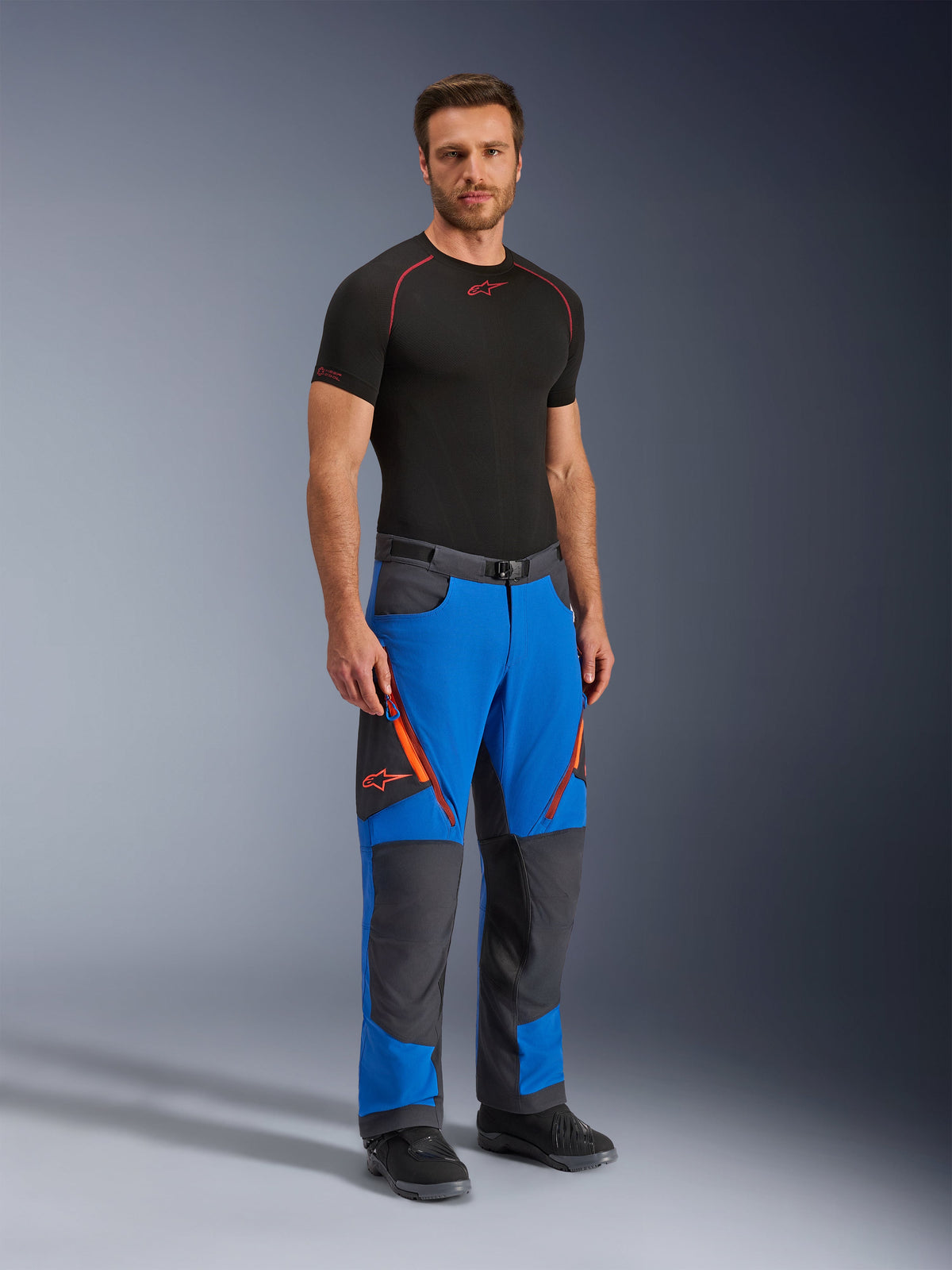 Alpinestars Maxdura Dual Hose, Adventure-Motorradhose, Royalblau und Dunkelgrau, mit orangefarbenen Reißverschluss-Akzenten und Logos, getragen von einem Modell mit schwarzem Base Layer und Stiefeln, technisches Design mit verstärkten Paneelen für Dual-Sport-Fahrten