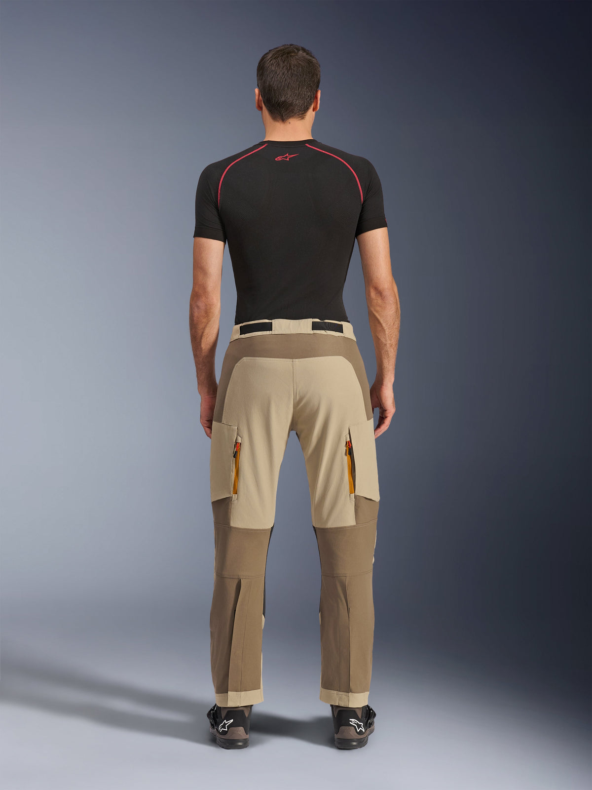 Alpinestars Maxdura Dual Pants in Walnut Aluminum, rear view showing two-tone brown and tan design, adventure riding pants with zippered cargo pockets and reinforced seat, worn with a black compression shirt and motorcycling boots.