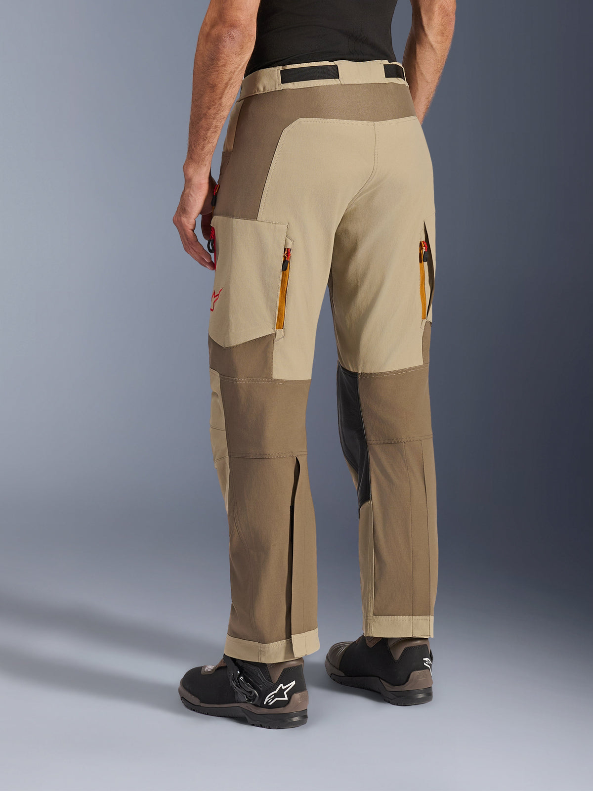 Alpinestars Maxdura Dual Pants in Walnut Aluminum, rear side view, adventure motorcycle pants featuring tan and brown panels, zippered rear exhaust vents, large cargo pockets, and over-the-boot design worn with black and brown boots