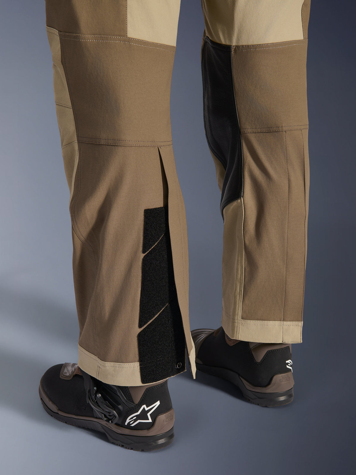 Alpinestars Maxdura Dual Pants in Walnut Aluminum, detailed view of the lower leg and hem featuring an over-the-boot design with adjustable hook-and-loop closures and reinforced inner panels, paired with black motorcycle boots.