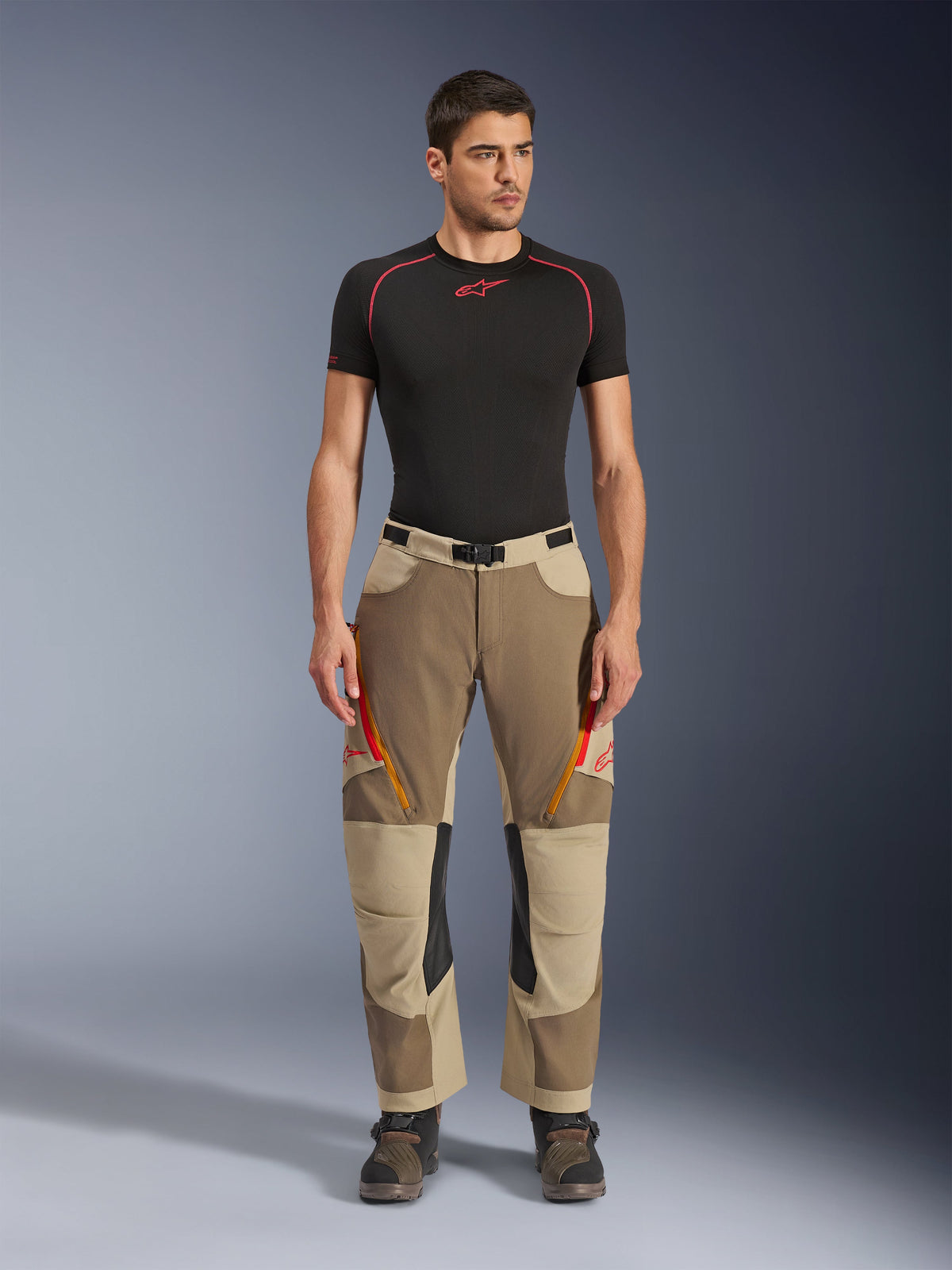 Alpinestars Maxdura Dual Pants, Adventure and Dual-Sport Riding Pants, Walnut Aluminum, worn with a black short-sleeve compression shirt and adventure boots, featuring orange zippered vents and red logos on the thighs, black waist belt with buckle, over-the-boot design for motorcycle riding.