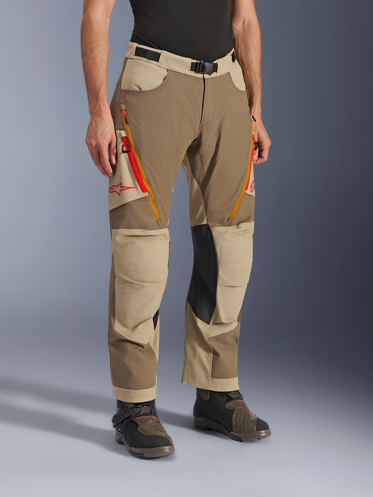 Alpinestars Maxdura Dual Pants, Walnut Aluminum brown and khaki adventure riding pants, featuring orange and red zippered thigh vents, reinforced black leather inner knee panels, and red Astars logo, worn with technical boots.