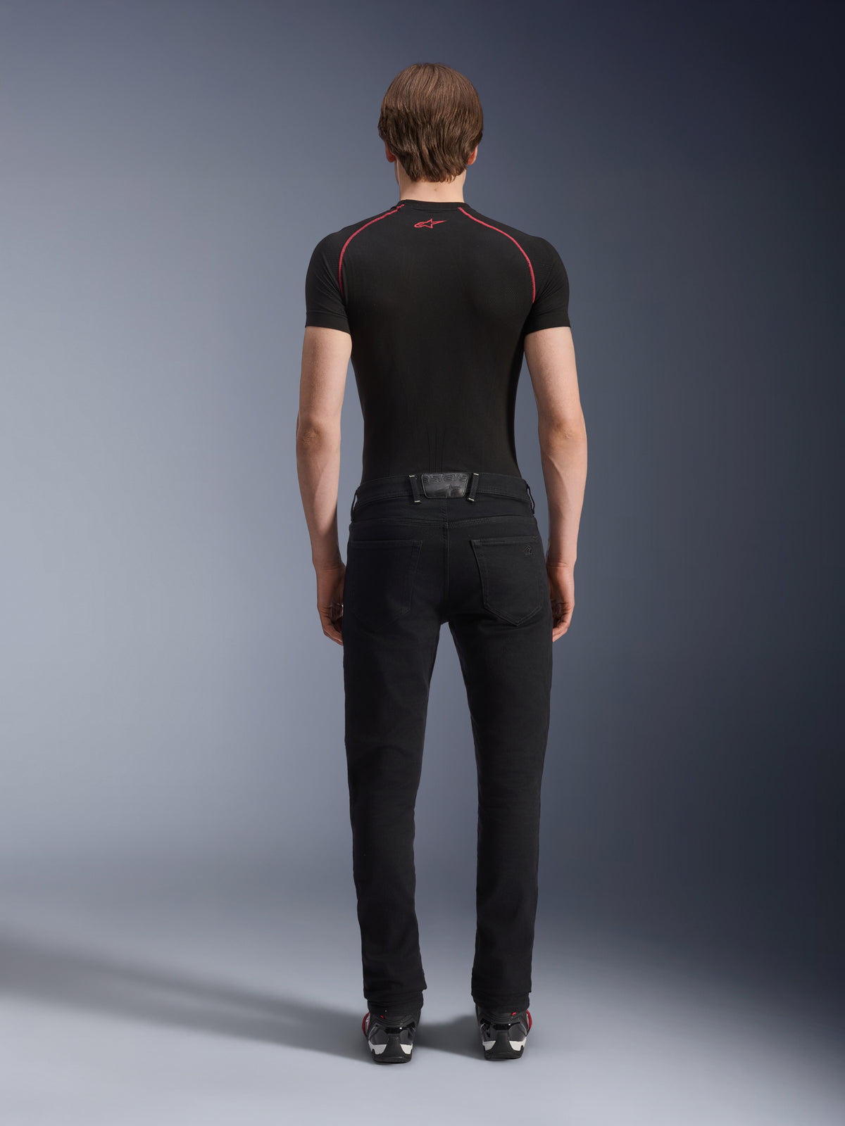 Alpinestars Copper V3 Jeanshose in Black Rinse, Rückansicht an einem Modell, reguläre Motorradjeans mit geradem Bein aus Stretch-Denim, kombiniert mit einem schwarzen kurzärmeligen Kompressionsshirt und Motorradschuhen, urbane Motorradbekleidung