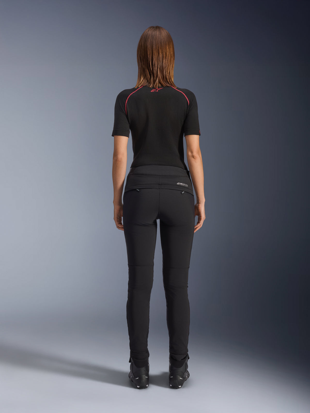 Alpinestars Alamosa Damenhose, Schwarz, Rückansicht mit schmaler Textilkonstruktion, Gesäßtaschen mit Reißverschluss und reflektierendem MO.ST.EQ.-Branding auf der Rückenpasse, getragen mit einer schwarzen kurzärmeligen Basisschicht und schwarzen Motorradstiefeln.