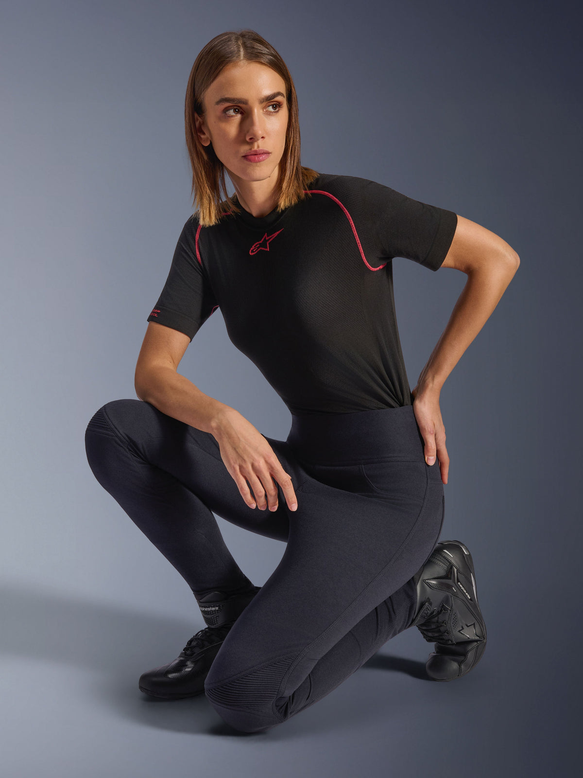 Frau in Alpinestars Stella Flex-AST Leggings, schwarze hoch taillierte Skinny-Motorrad-Leggings mit Akkordeon-Knieeinsätzen, kombiniert mit einem schwarzen kurzärmeligen Funktions-T-Shirt mit rosa Nähten und Motorradstiefeln.