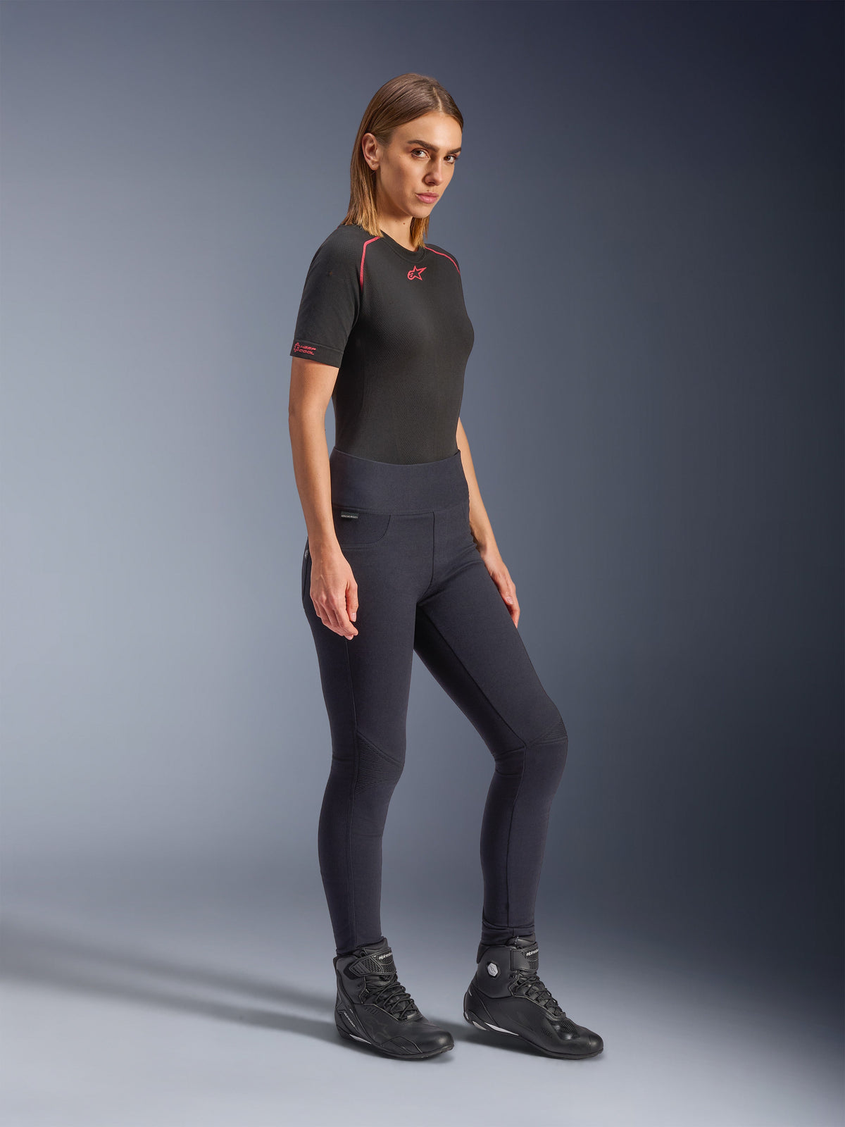 Model trägt Alpinestars Stella Flex-AST Damen-Leggings in Schwarz, hoch taillierte Skinny-Passform mit Akkordeon-Kniepaneelen, kombiniert mit einem schwarzen kurzärmeligen Alpinestars-Oberteil und schwarzen Motorradstiefeln, Ganzkörper-Studioaufnahme.