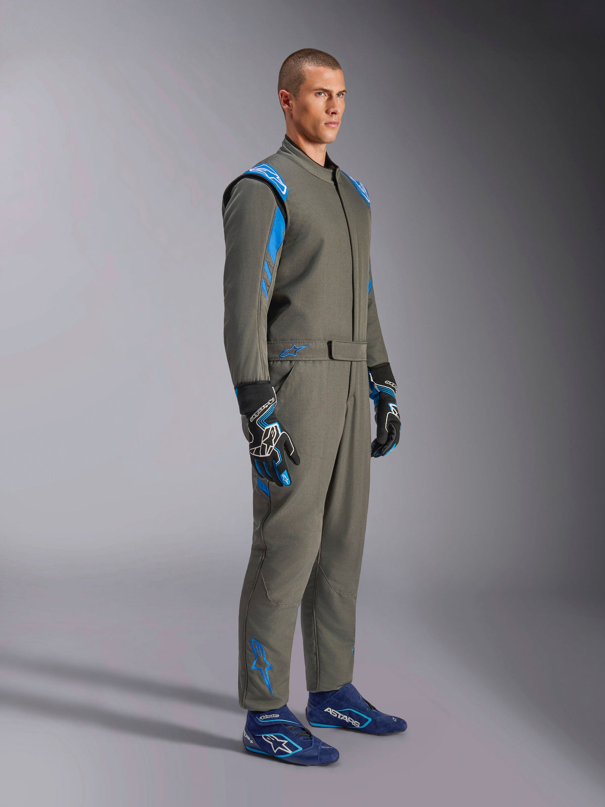 Alpinestars GP Race V3 Suit FIA in Anthracite Blue, one-piece auto racing suit worn by a male model, features bright blue accents on the shoulders and ankles, paired with coordinating racing gloves and blue racing shoes, professional motorsport gear.