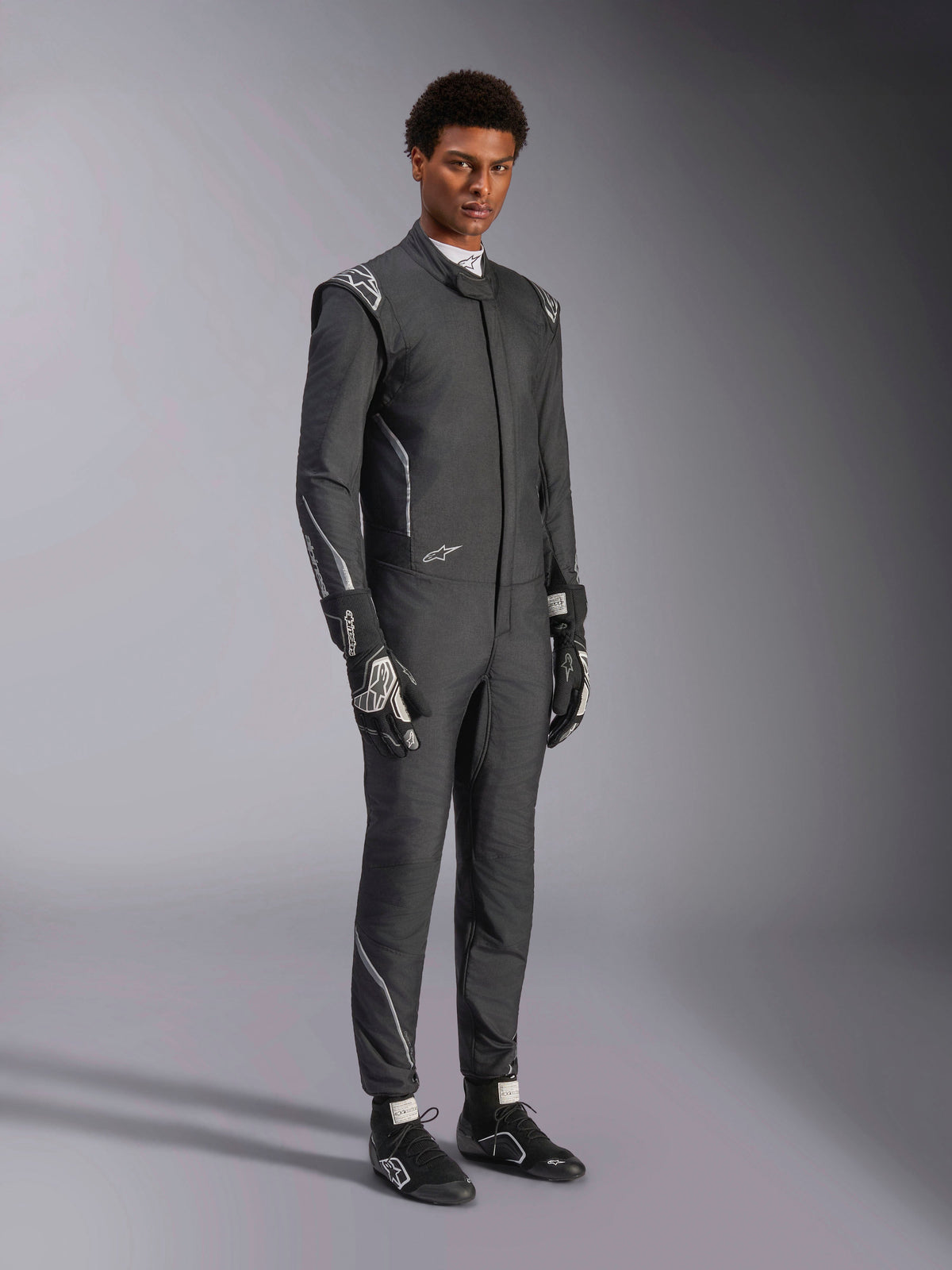 Alpinestars Hypertech V3 Suit, Auto Racing Suit, Black and Dark Shadow, worn by male model with matching black gloves and racing shoes, athletic slim fit, features Alpinestars logos and light gray accents, professional motorsport protective gear