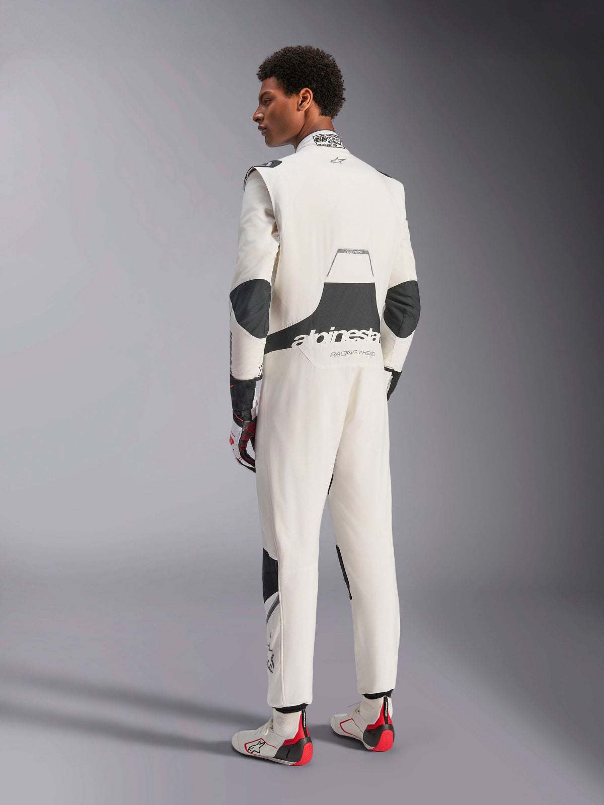 Alpinestars Hypertech V3 Anzug, professioneller Autorennanzug, Weiß, Schwarz und Silber, Rückansicht des Models mit schwarzen Stretcheinsätzen im Lendenbereich und an den Ellbogen, Alpinestars-Branding auf dem Rücken, kombiniert mit passenden Rennhandschuhen und Stiefeln für einen kompletten Motorsport-Look