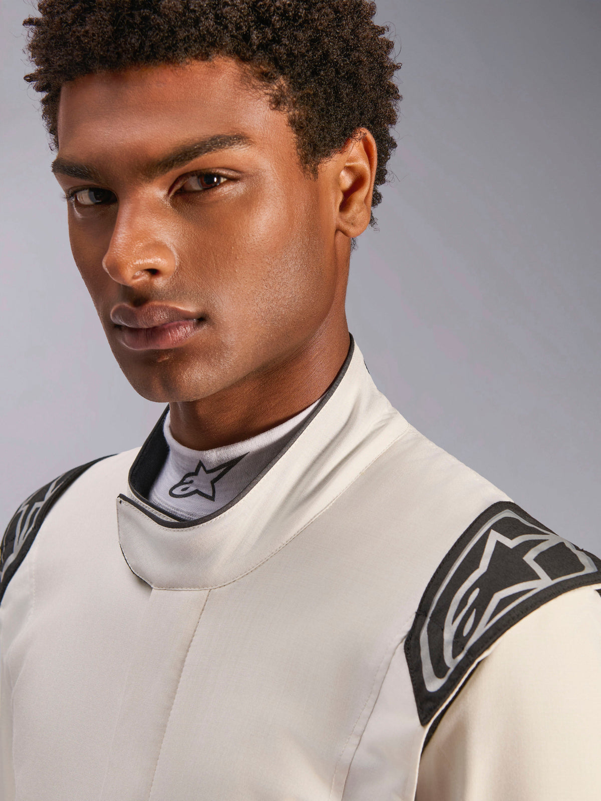Nahaufnahme eines männlichen Models im Alpinestars Hypertech V3 Anzug in Weiß Schwarz Silber, mit hohem Kragen mit sicherem Verschluss und Alpinestars-Logos auf den Schultern, professionelle Rennsportbekleidung.